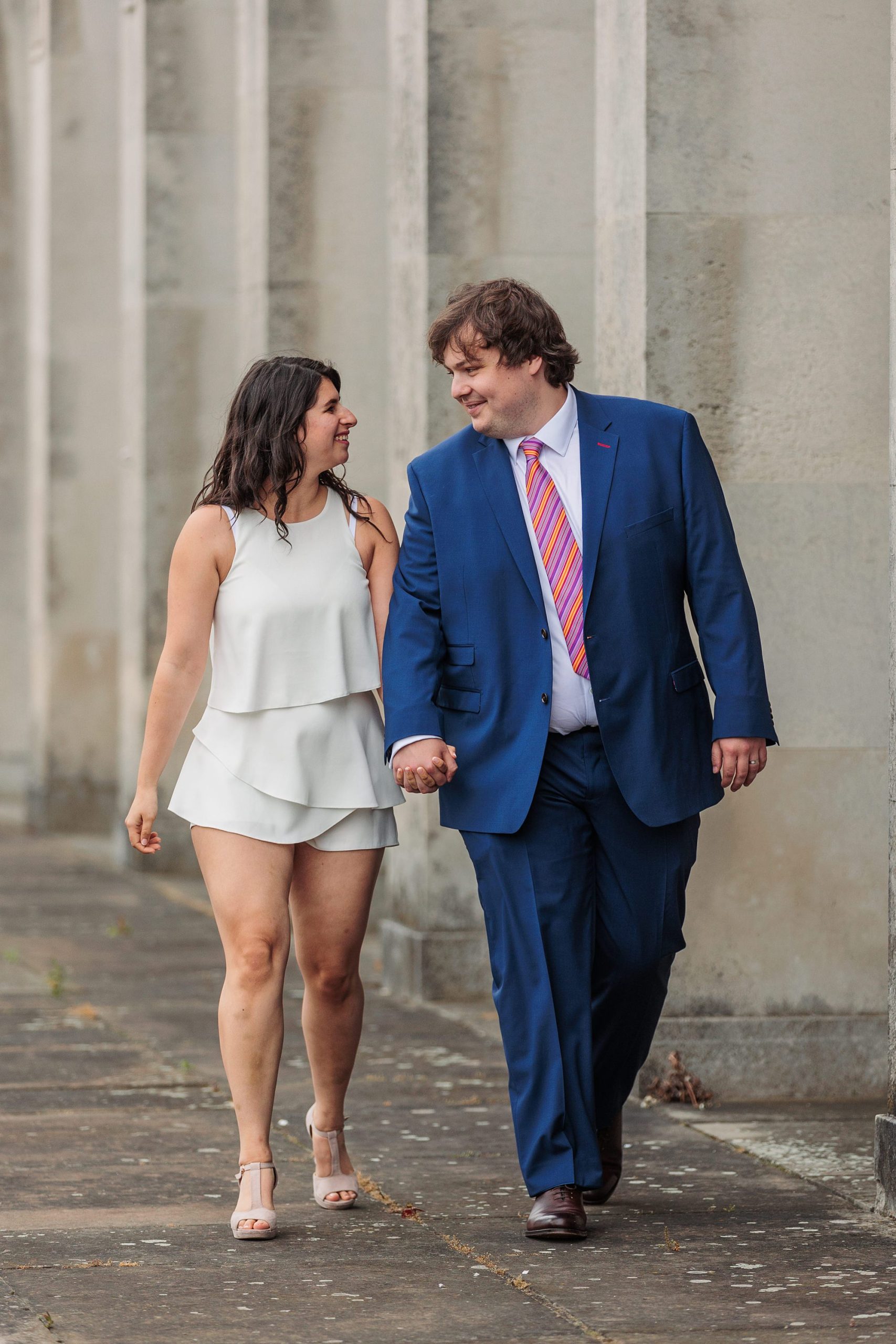 A man in a blue suit with a colorful striped tie holds hands with a woman in a white sleeveless dress. They smile at each other as they walk along the stone walkway outside Hertford Register Office, savoring the joy of their special day.