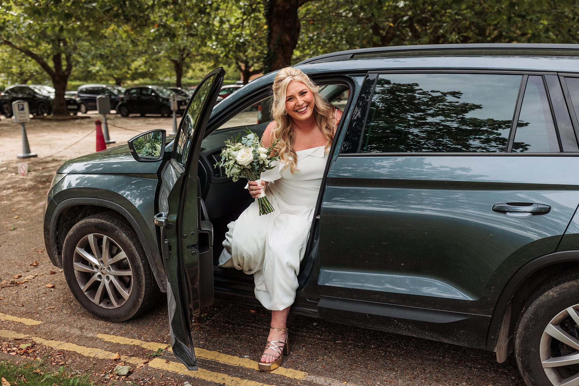 A bride in a white dress steps out of a dark green car, bouquet in hand, her smile radiant against the backdrop of trees and parked cars near Hertford Register Office.