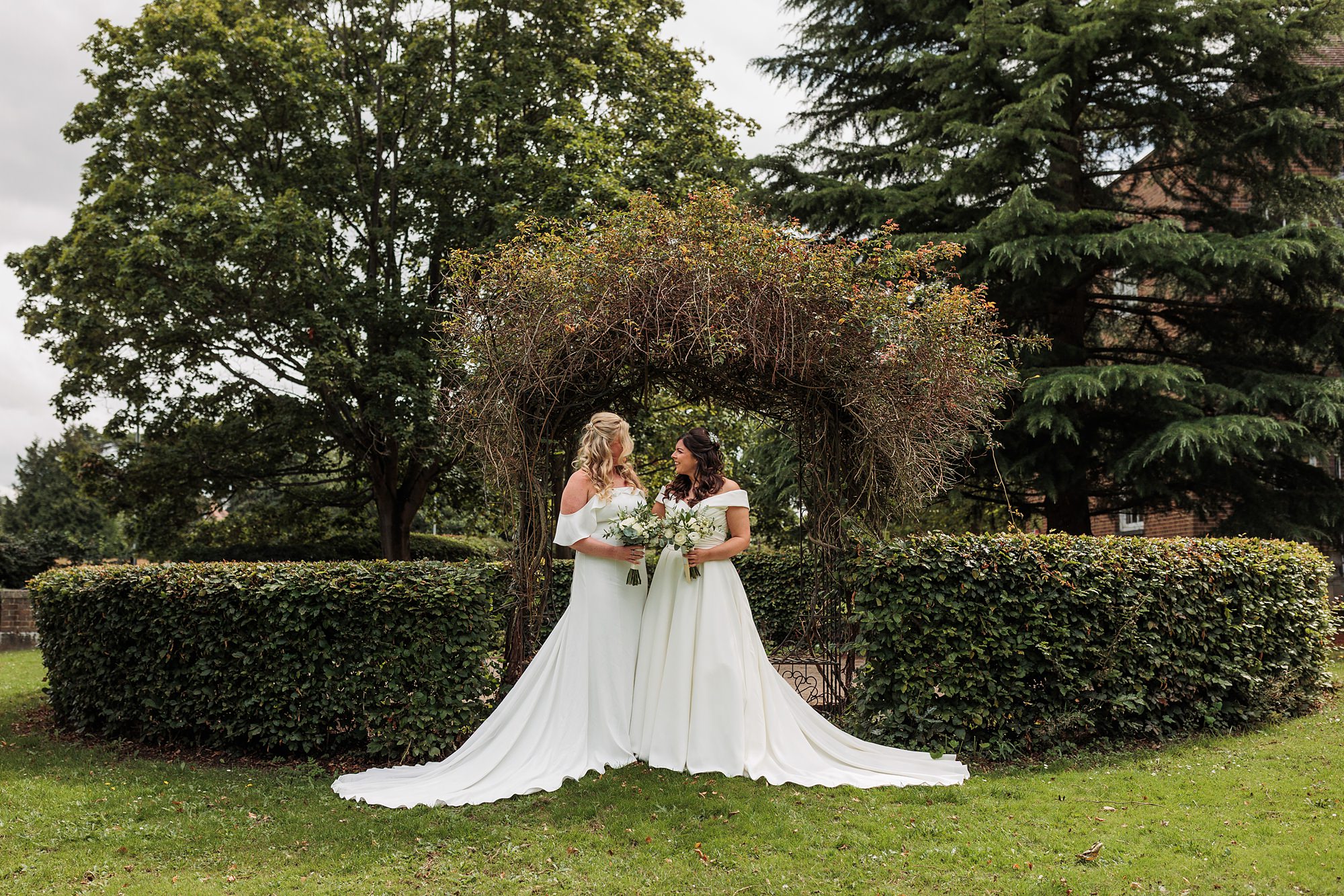 Two brides in white dresses stand together before a greenery-covered arch in a garden, reminiscent of the charm found at Hertford Register Office. They hold bouquets and are surrounded by lush trees and bushes, smiling and gazing at each other lovingly.