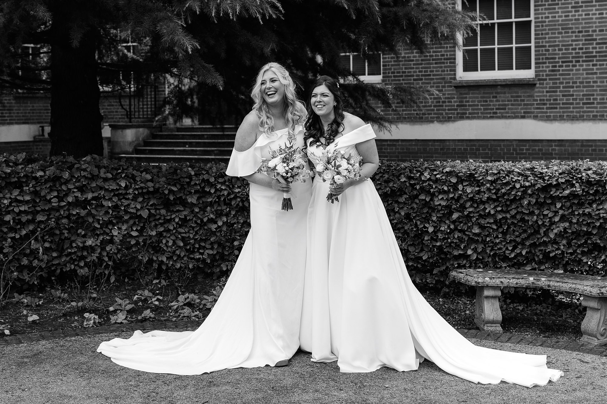 Two women in white wedding dresses stand outdoors at the Hertford Register Office, smiling and holding bouquets. They are surrounded by greenery with a brick building in the background. Their dresses have long trains, and they appear joyful.