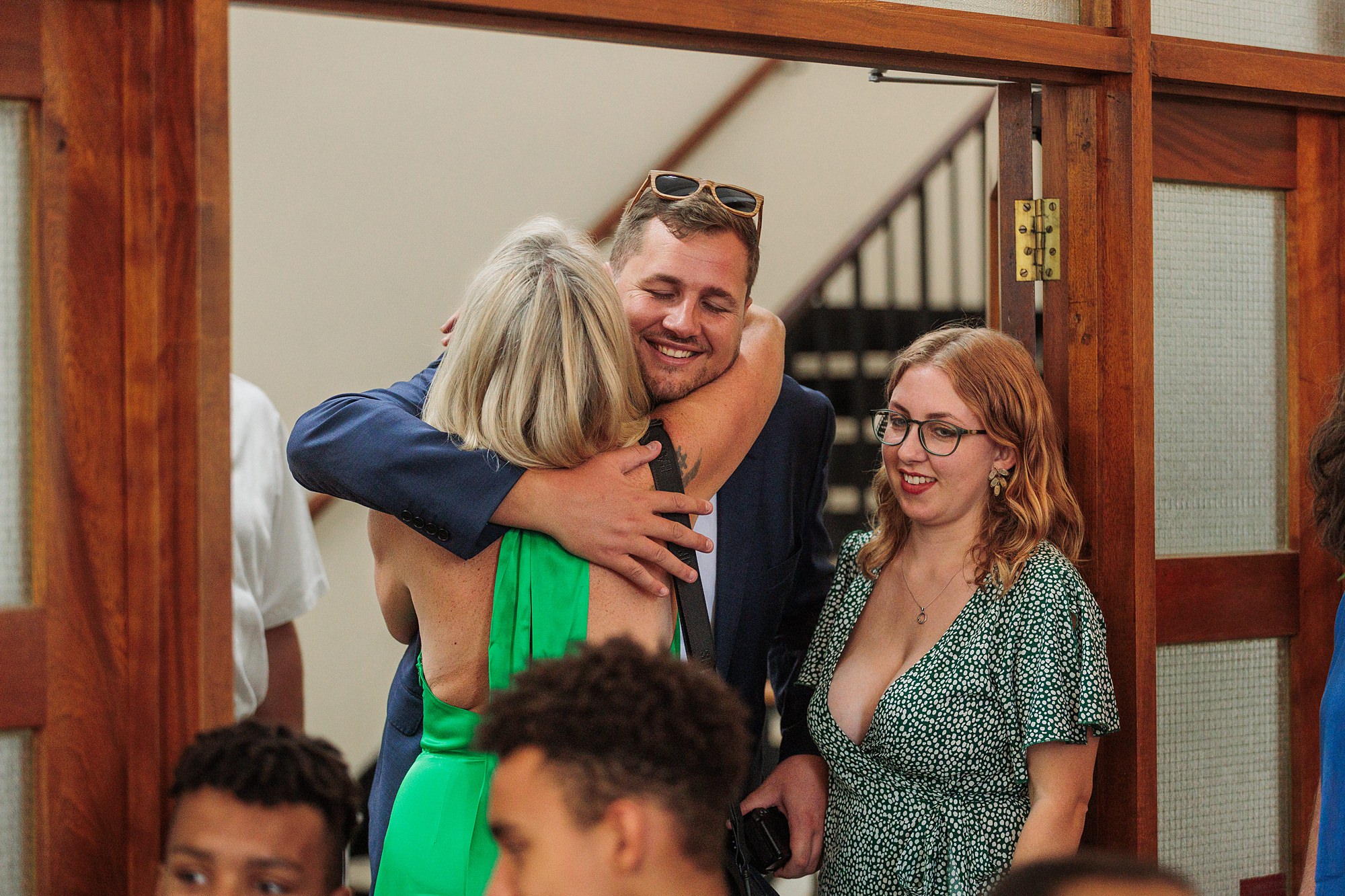 A man in a blue suit warmly embraces a woman in a green dress at the Hertford Register Office. Nearby, another woman in glasses and a green floral dress smiles, as a staircase looms in the background. Others are partially visible, adding to the cozy indoor gathering.
