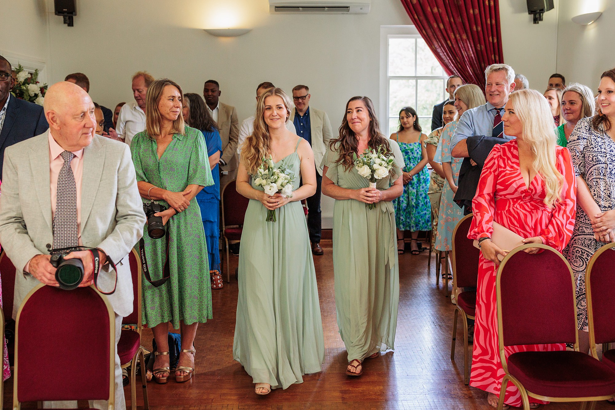 Two bridesmaids in light green dresses walk down the aisle at Hertford Register Office, holding bouquets. Guests seated in rows on either side watch, some with cameras. The room features red curtains and wooden floors, adding a touch of elegance to the ceremony.