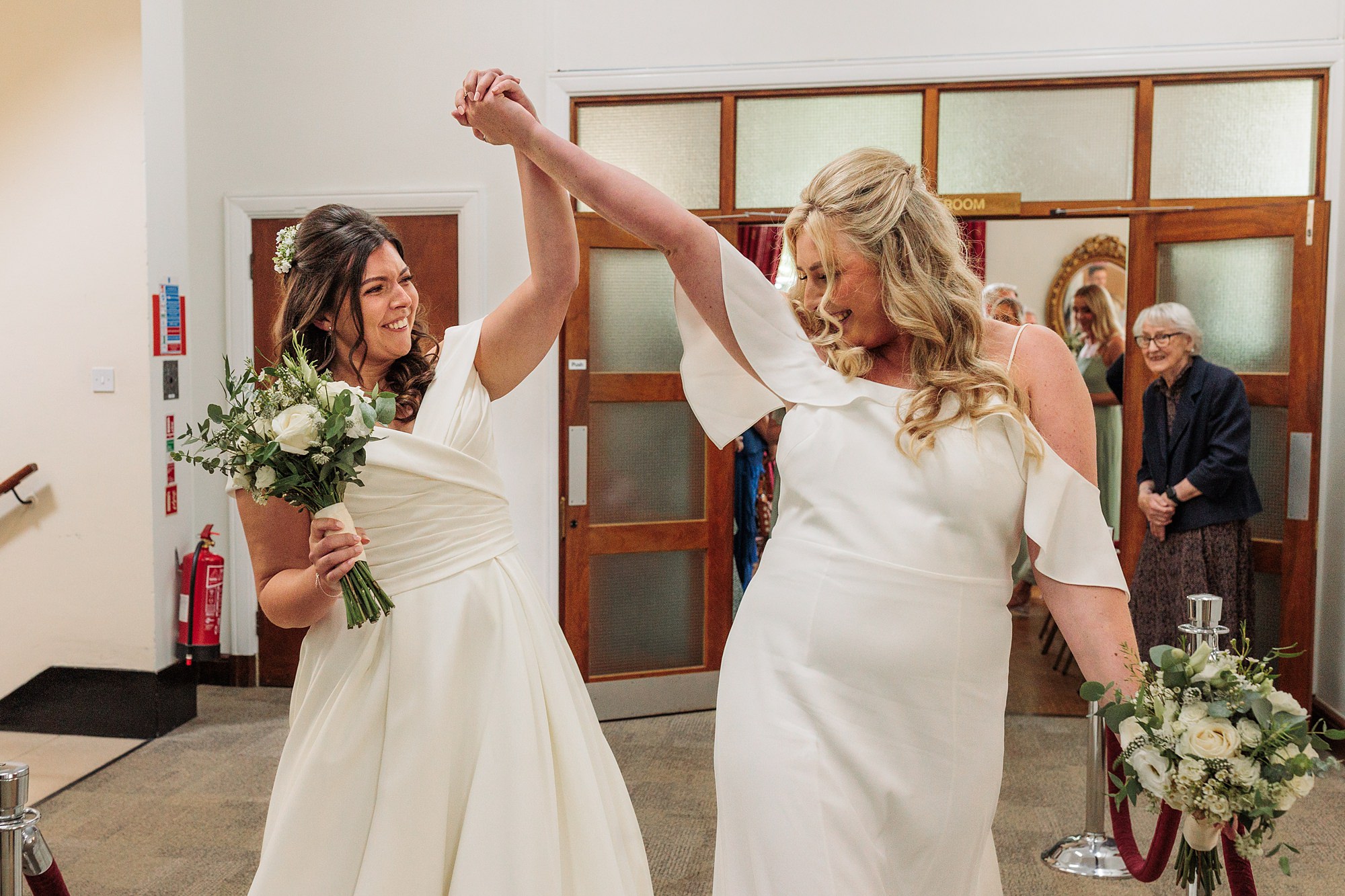 Two brides in white gowns joyfully dance and hold hands in the hallway of Hertford Register Office, each holding a bouquet. A woman in the background smiles while watching them, amid closed wooden doors.