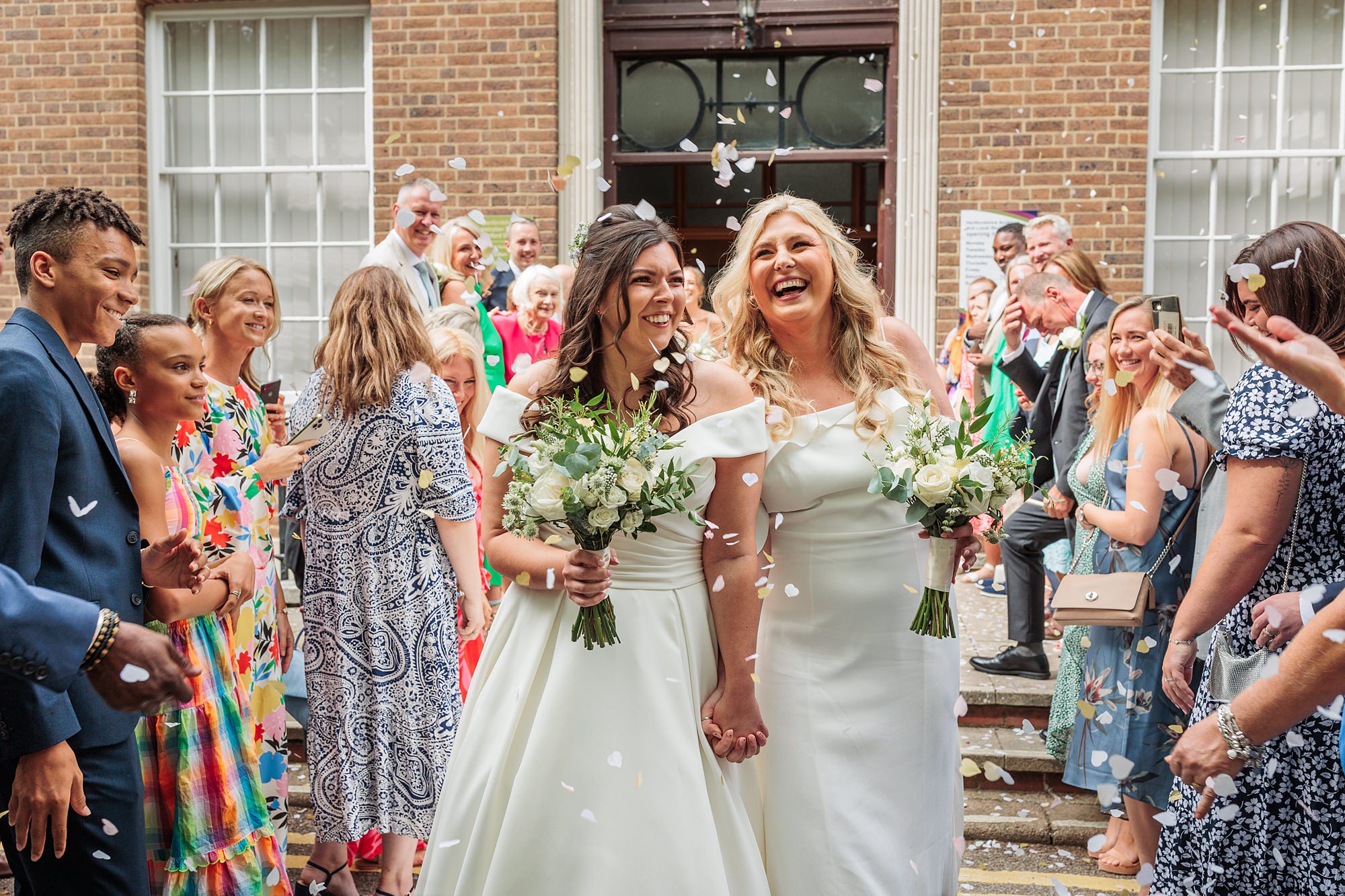 Two brides in white dresses, each clutching a bouquet, walk hand in hand and smile joyfully. Their guests celebrate by tossing flower petals at Hertford Register Office, where the large windows frame this outdoor celebration of love.