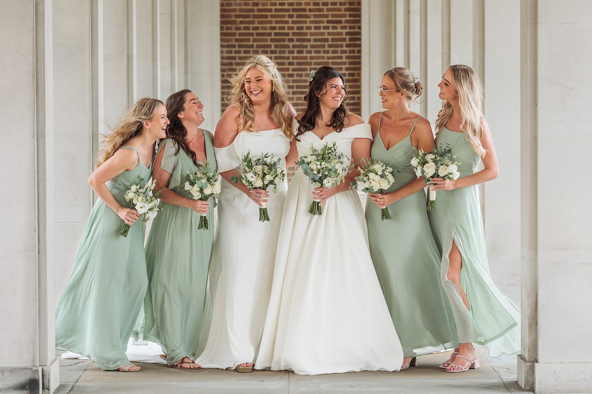 Six women in pastel green dresses pose joyfully with a bride in a white gown at the Hertford Register Office. They stand under an archway, holding bouquets of white flowers, laughing and smiling against a backdrop of cream pillars and a brick wall.
