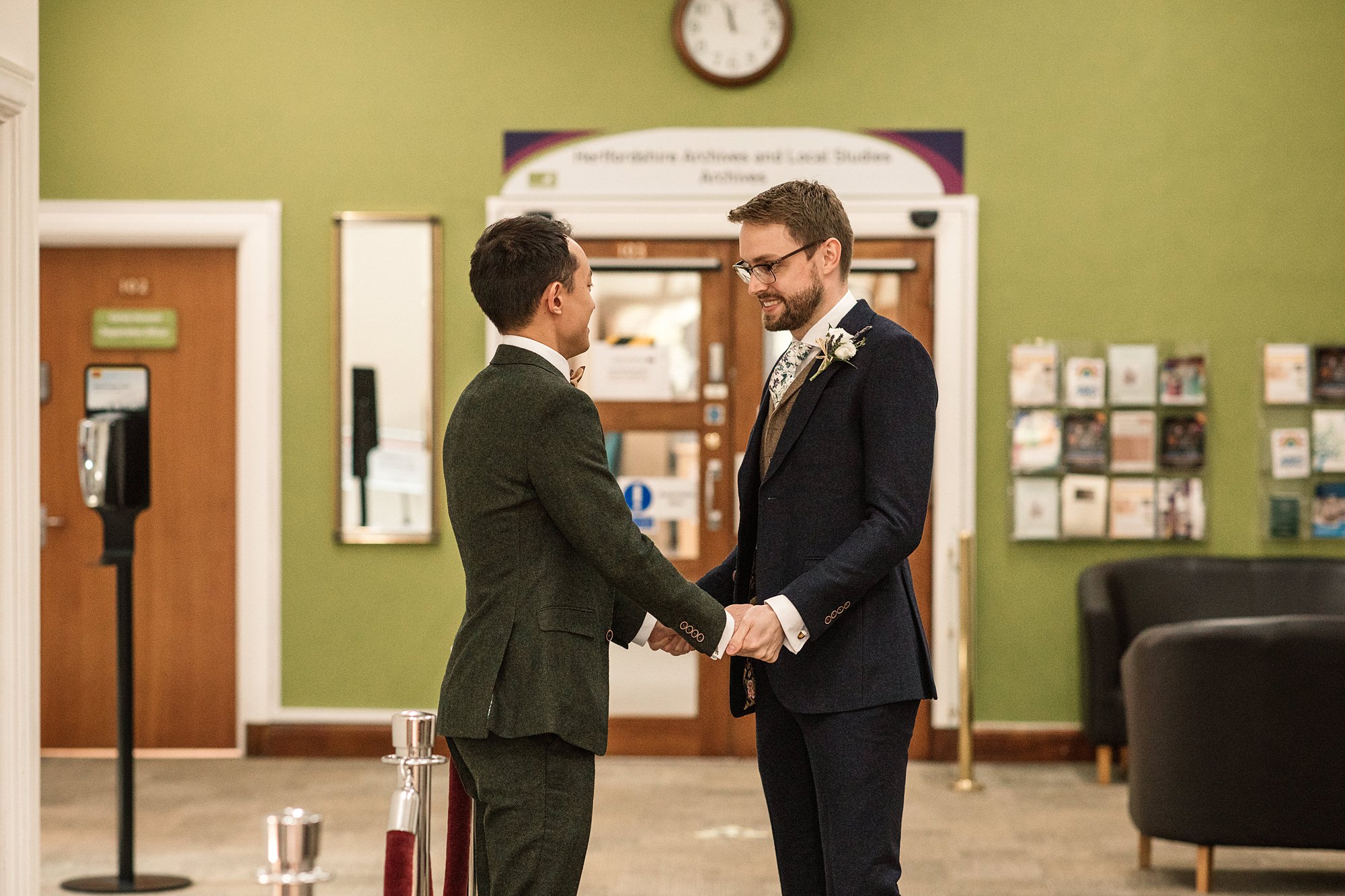 Two men in formal suits stand holding hands and smiling at each other in a room at the Hertford Register Office, with a green wall and a clock above a double doorway. Chairs and brochures are visible in the background.