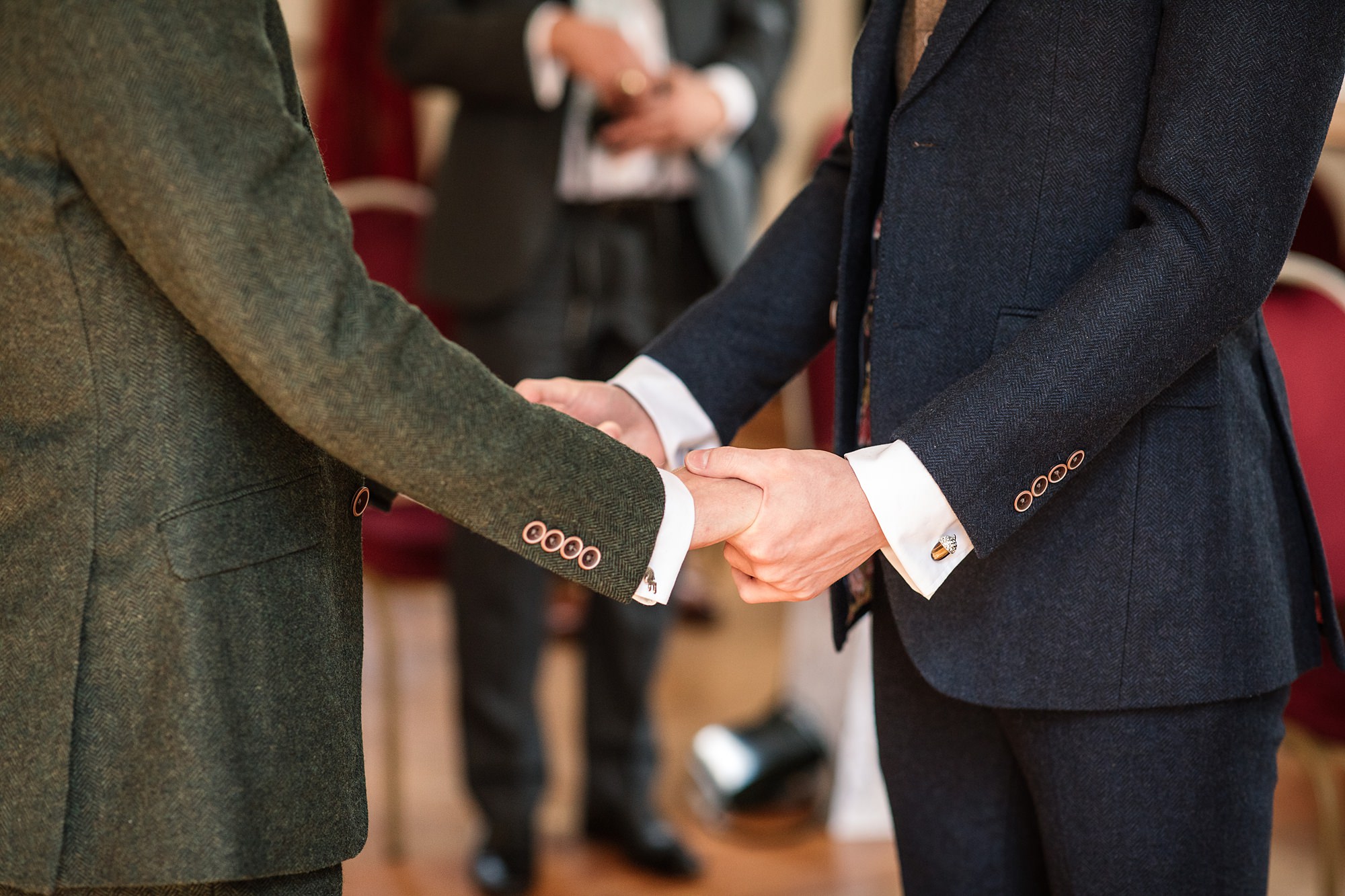 In a ceremony at the Hertford Register Office, two people in formal suits hold hands. Their faces remain unseen; one dons a green suit, the other dark blue. In the blurred background, another suited figure stands with something in hand.