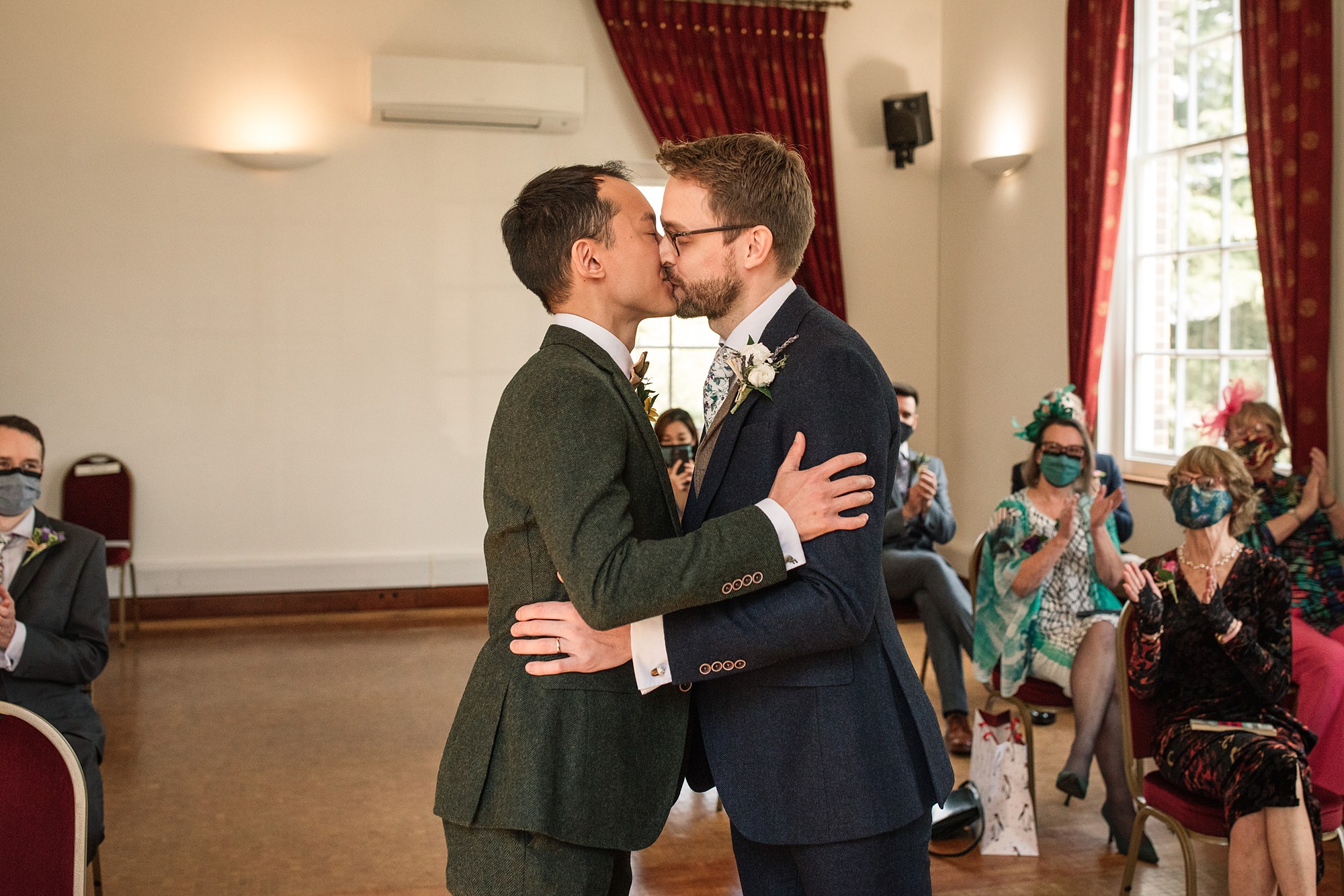 In a warmly lit room with red curtains and large windows at Hertford Register Office, two grooms kiss, dressed in formal suits—one in dark green and the other in navy. They are surrounded by seated guests, some wearing masks, sharing the moment with joy.