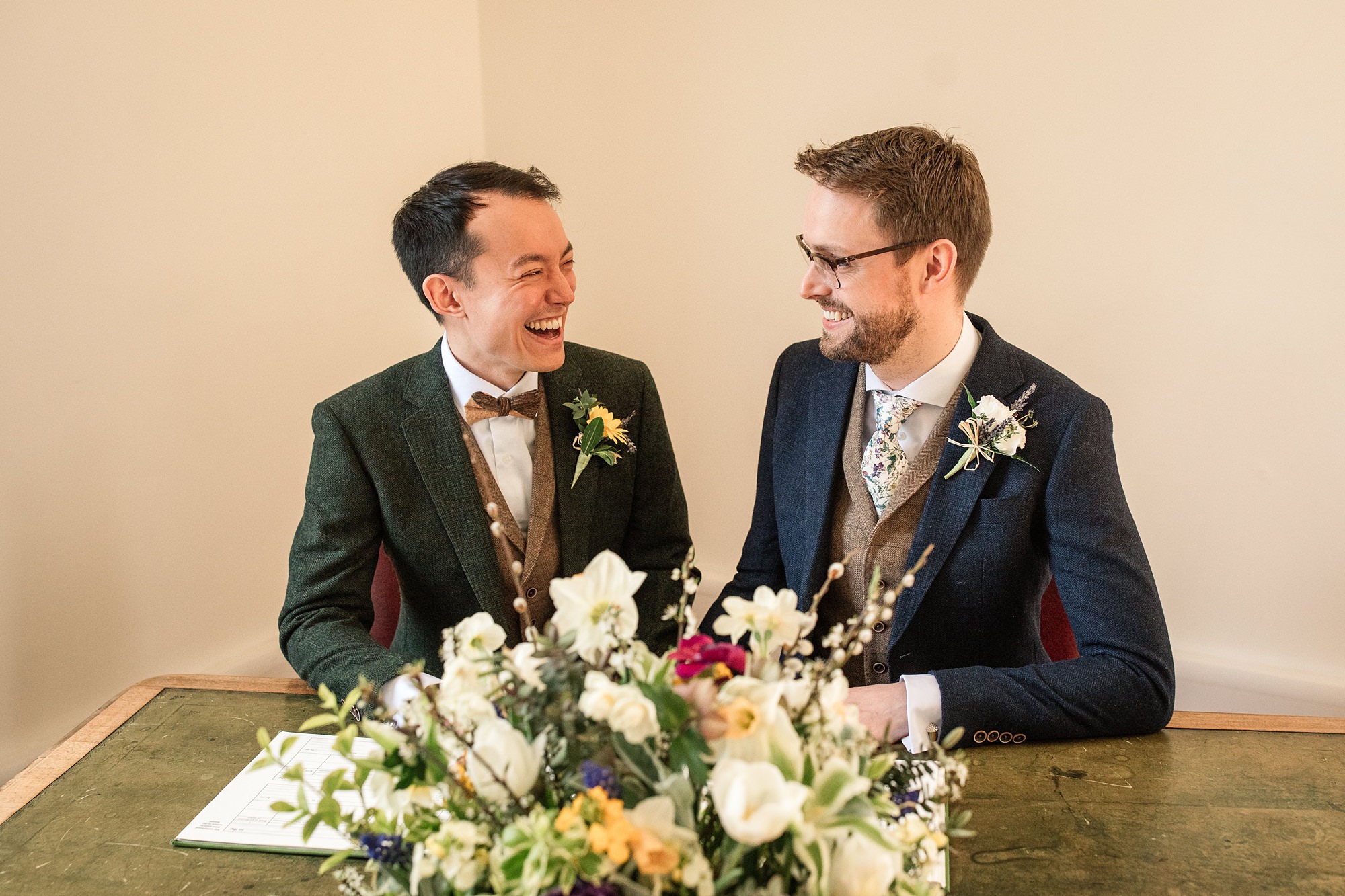 Two people in suits smile at each other while seated at a table adorned with a vibrant floral arrangement. Both wear boutonnieres, and the warmly lit setting at Hertford Register Office suggests a wedding or formal event.
