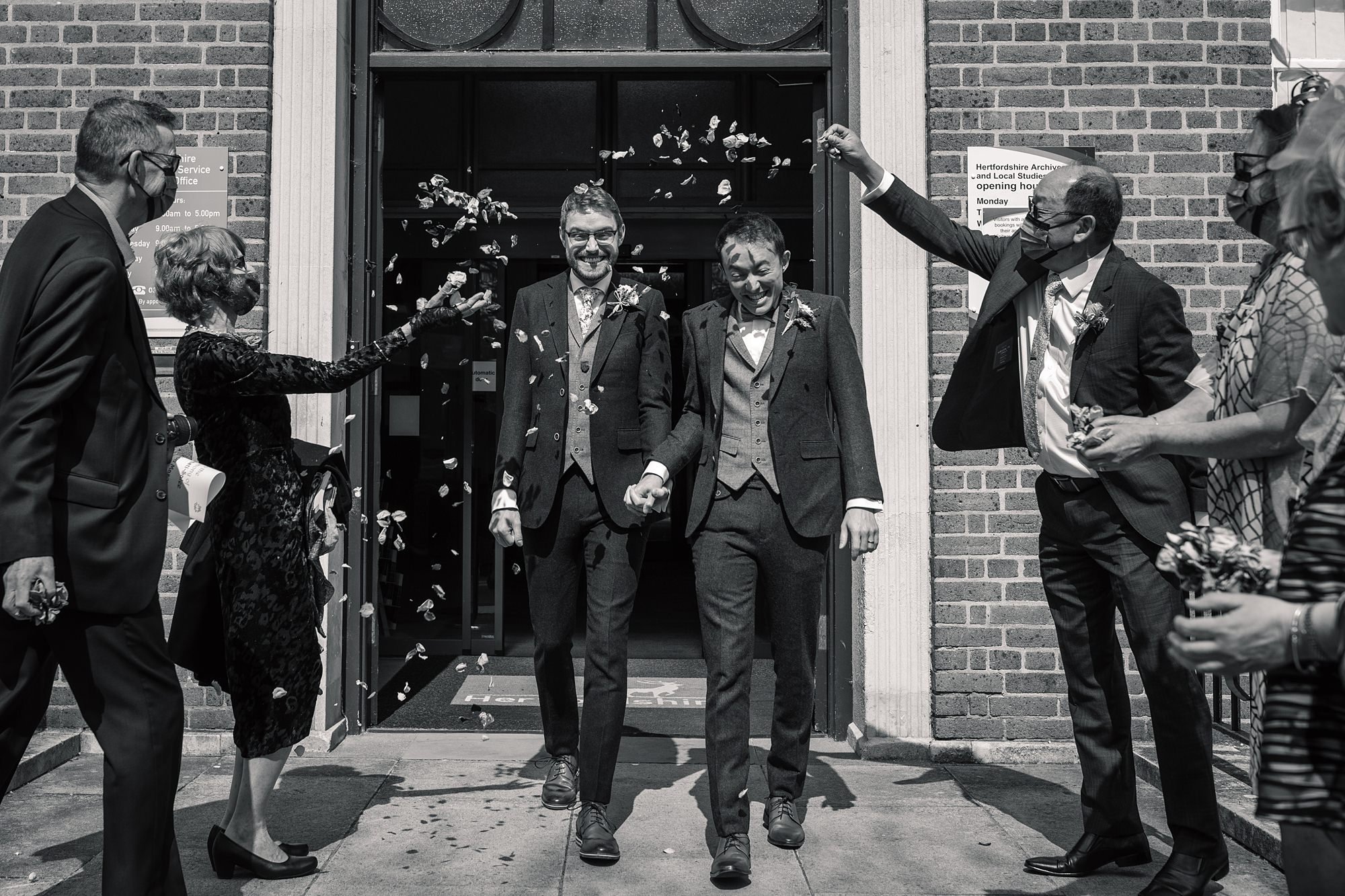 Two grooms holding hands walk out of Hertford Register Office, smiling as they are showered with confetti by well-dressed people on either side. Some individuals are wearing masks. The scene is joyful and celebratory.