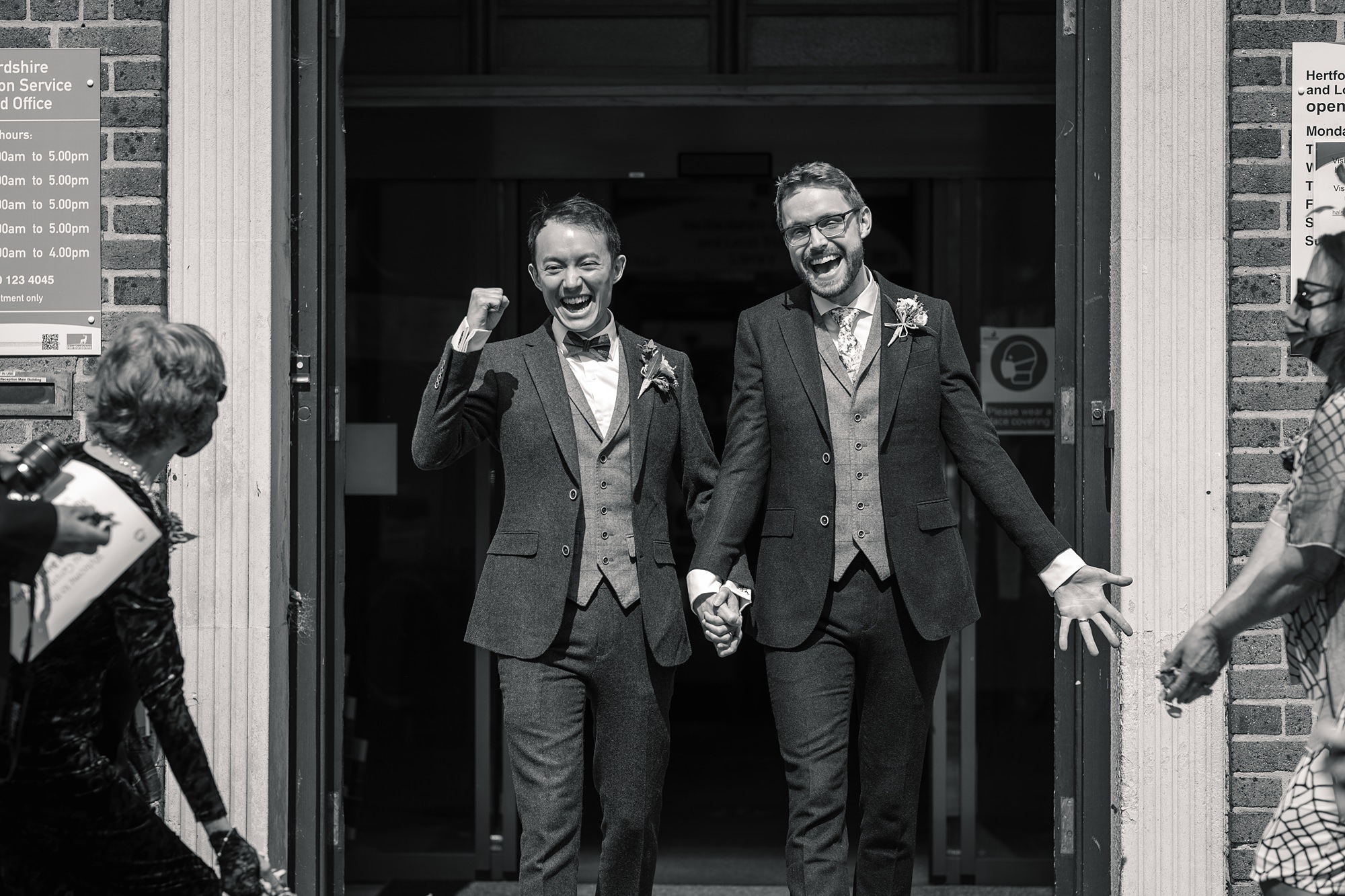 Two grooms in suits exit Hertford Register Office, holding hands and smiling joyfully. One raises a fist in celebration as people cheer and throw confetti. A sign with opening hours is visible on the wall, enhancing the festive, celebratory mood of this special moment.