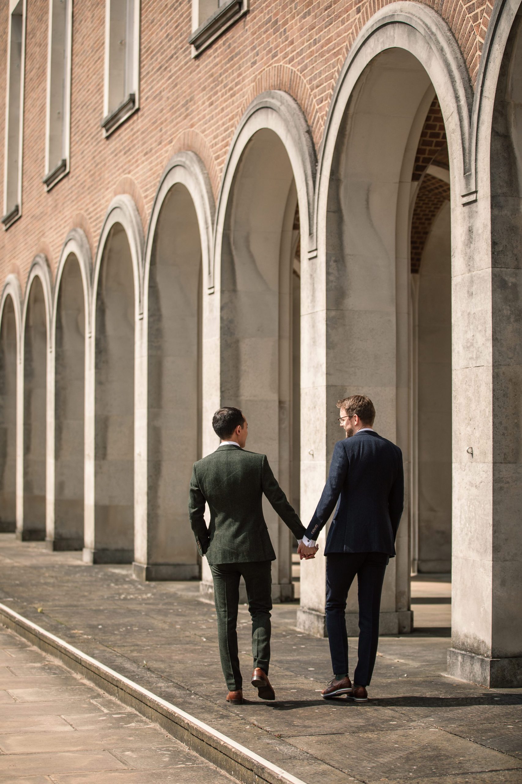 Two people in suits hold hands while walking along the pathway of the Hertford Register Office. They are under a row of arched columns on a brick building facade, one in a dark green suit and the other in dark blue.