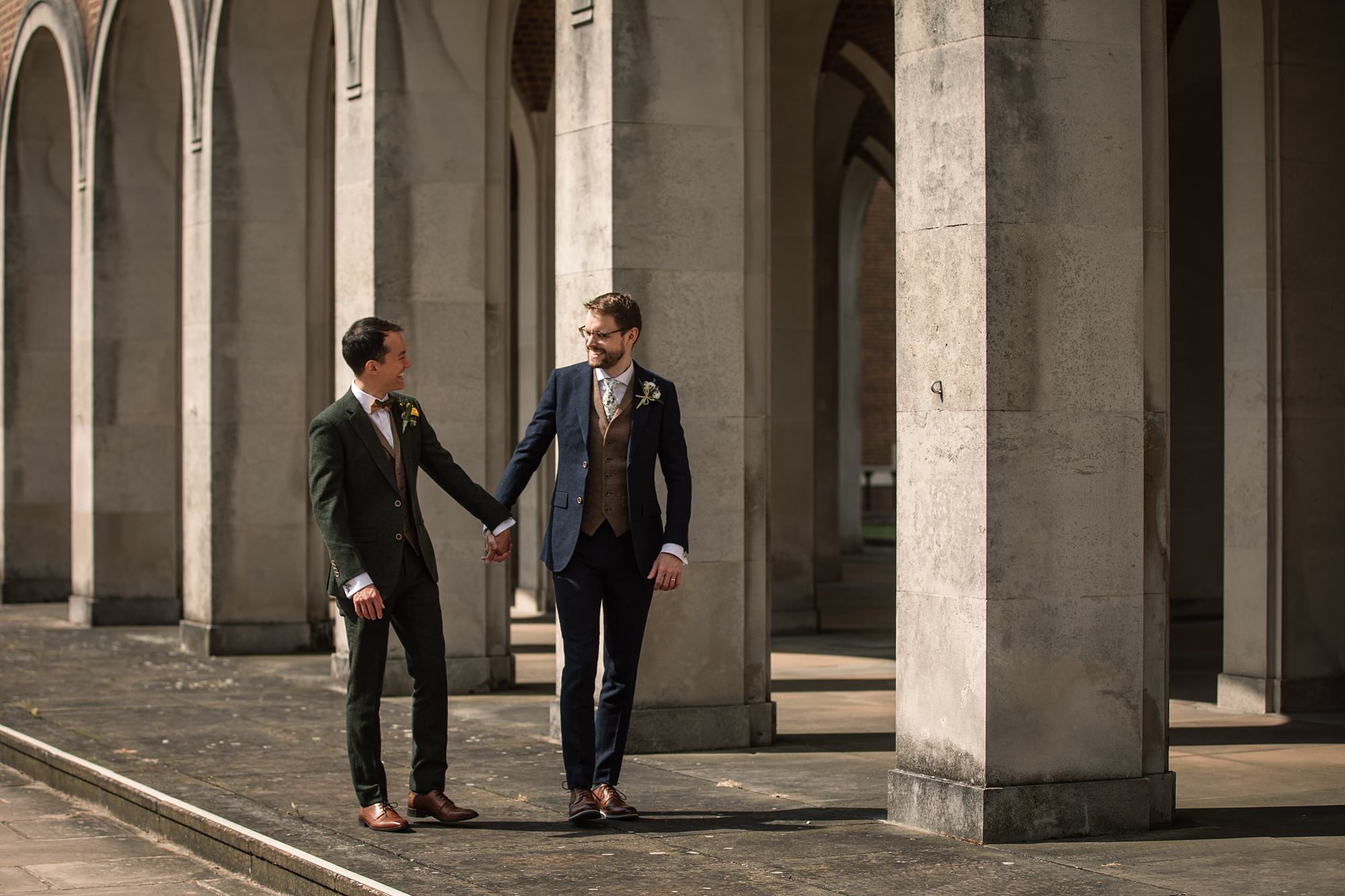 Two men in formal suits hold hands while walking under the stone archways of Hertford Register Office. The man on the left wears a dark green suit, and the one on the right sports a navy suit with a brown vest. They smile at each other, surrounded by the majestic columns.