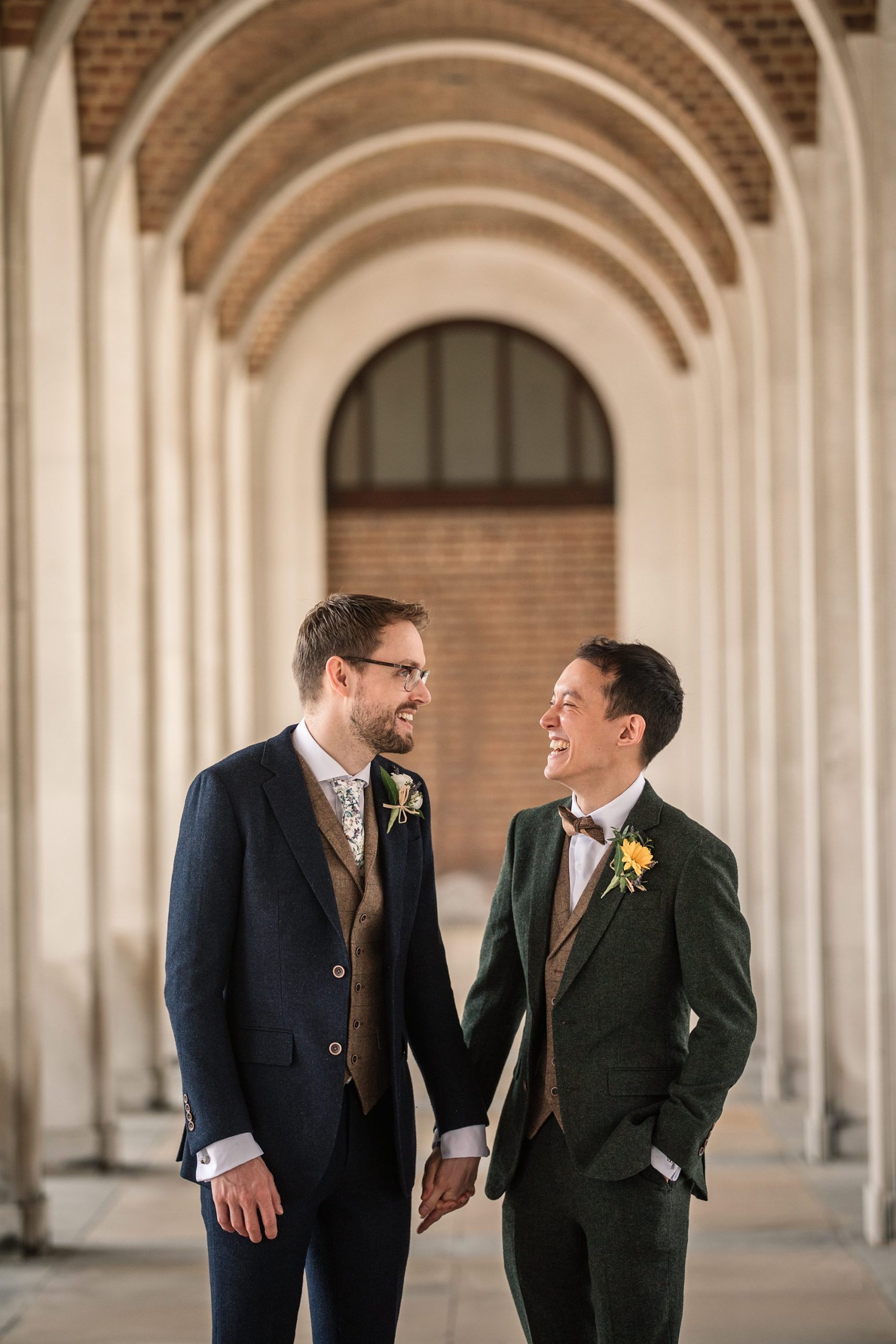 Beneath the elegant arches of the Hertford Register Office, two people in suits stand hand in hand, smiling at each other. One wears a navy suit with a floral tie and boutonniere; the other, a dark green suit with a yellow boutonniere. The backdrop boasts stunning brick and stone architecture.