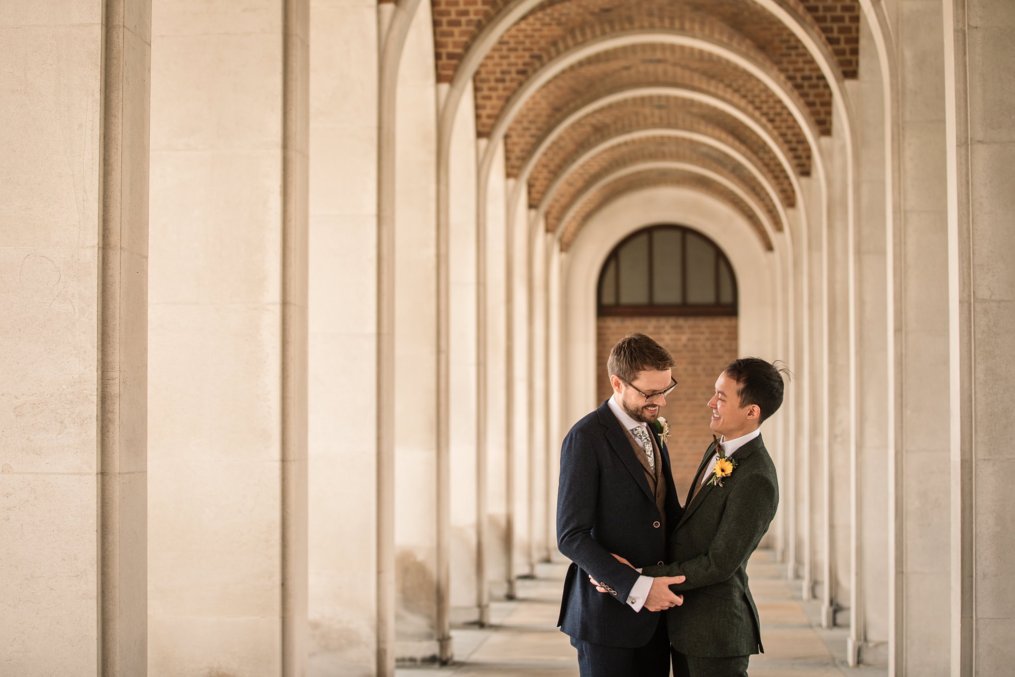 Two people in suits stand closely, smiling and embracing under a series of stone arches at the elegant and historic Hertford Register Office. One wears a blue suit with a tie, and the other a dark green suit adorned with a floral boutonniere.