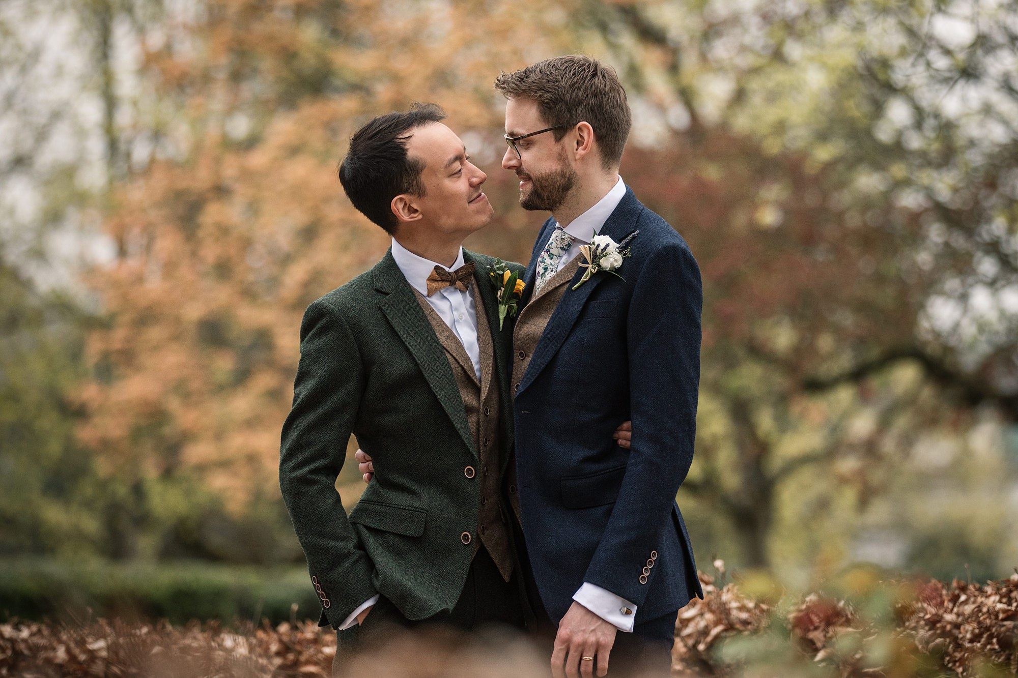 Two men in suits embrace outdoors near Hertford Register Office, surrounded by trees with autumn foliage. One wears a green suit, the other a dark blue suit. Both have flowers on their lapels and smile warmly at each other.