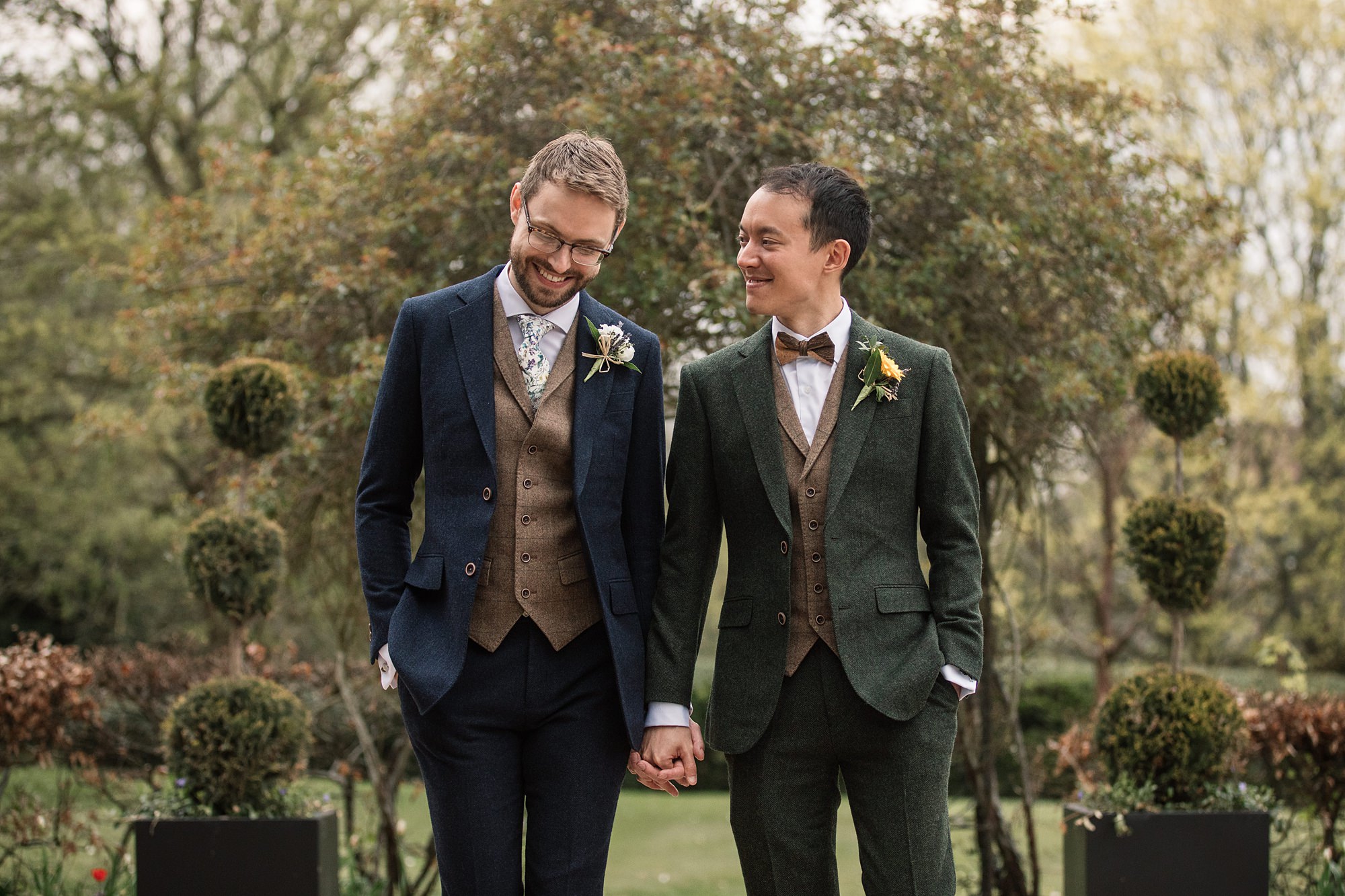 Two men in suits hold hands and smile at each other while walking outdoors, perhaps just after a visit to the Hertford Register Office. One wears a navy suit with a brown vest, the other a green suit with matching vest. Both have boutonnières, and trees are visible in the background.