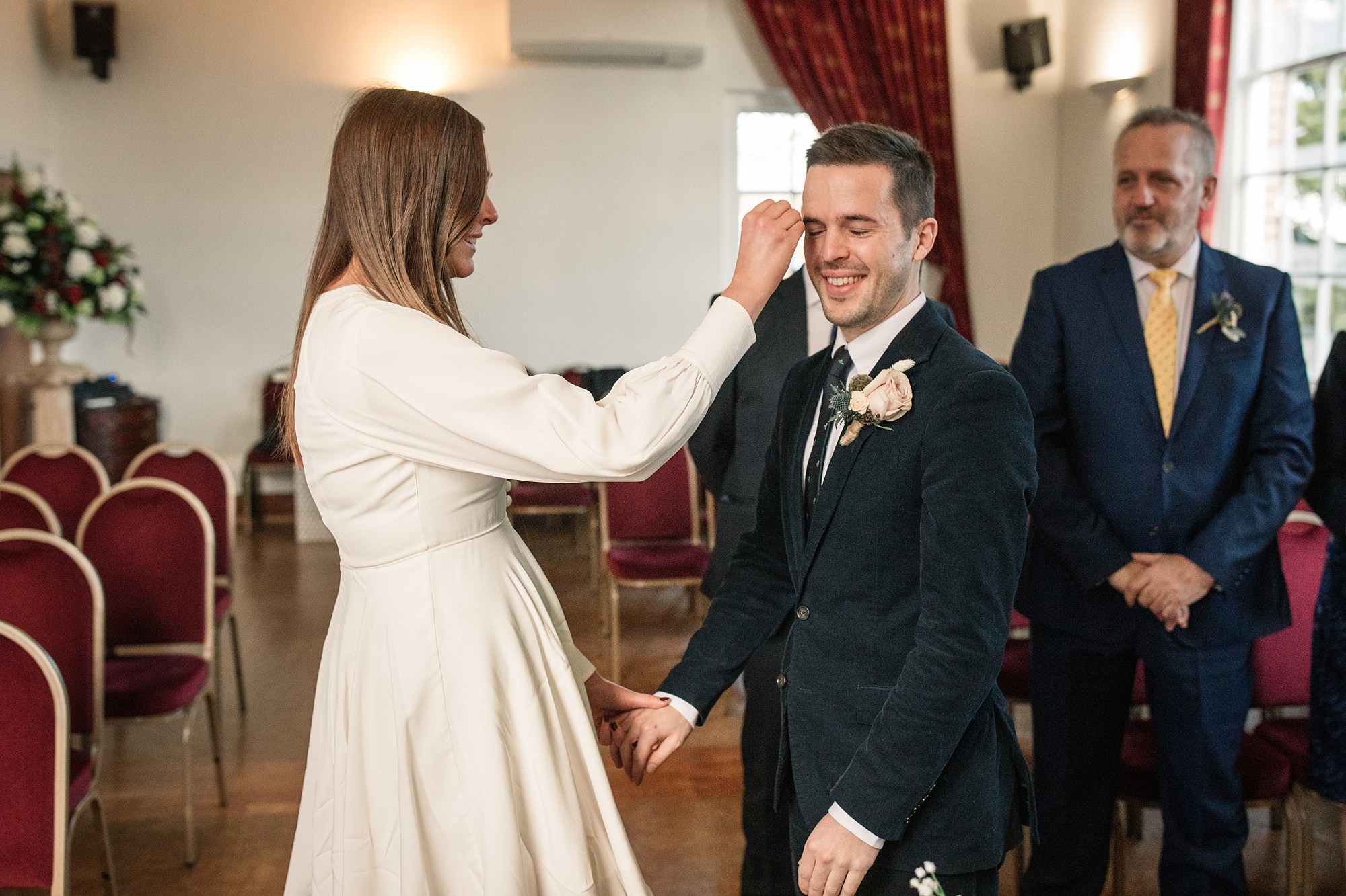 In Hertford Register Office, a woman in a white dress gently wipes the face of a smiling man in a suit, holding his hand. They are surrounded by red chairs and floral arrangements, while another suited man watches from the background with a smile.