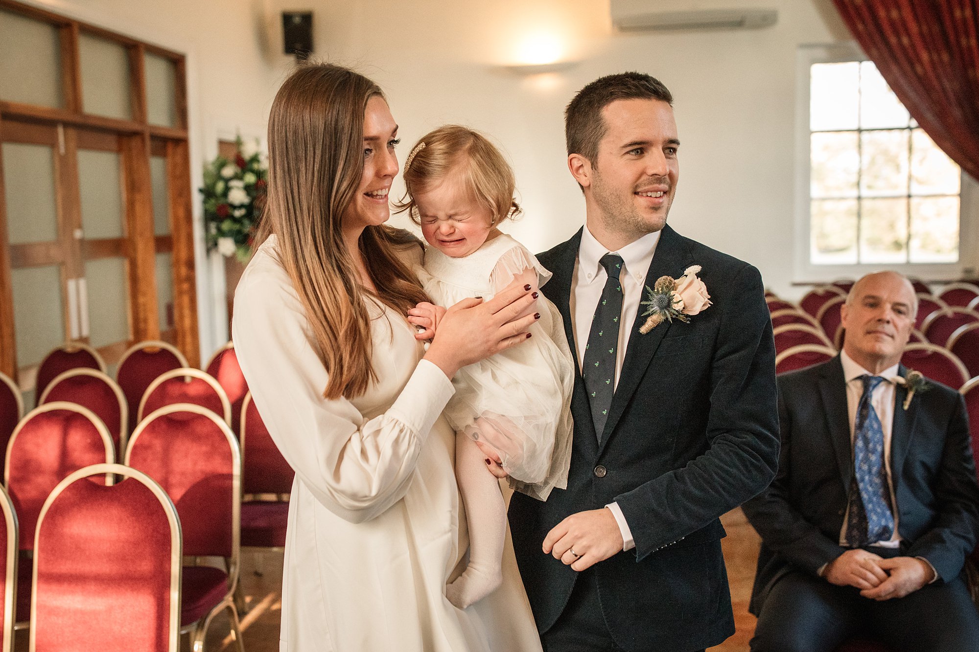 A woman and man, both smiling, hold a crying baby dressed in a white outfit at Hertford Register Office. They stand in a room with red chairs and large windows, while another man sits in the background, observing them.