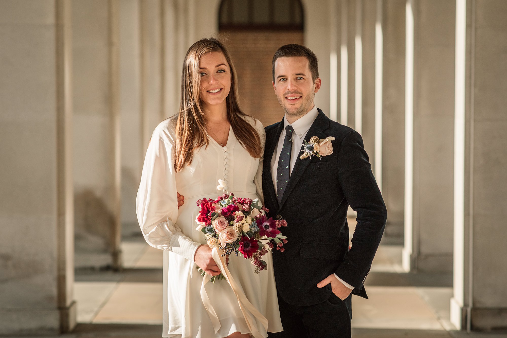 A couple posing happily in a sunlit corridor at Hertford Register Office. The woman wears a white dress and holds a bouquet of flowers, while the man is in a dark suit with a flower on his lapel. Both are smiling at the camera.