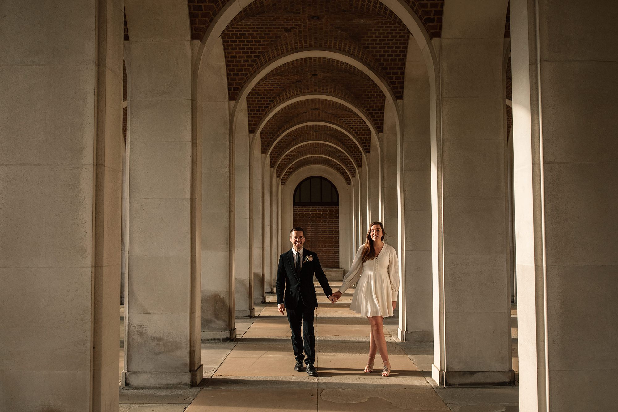 A couple walks hand in hand through the beautifully lit corridor of the Hertford Register Office, with tall stone arches embracing them. The man is in a dark suit and the woman in a white dress, both smiling as sunlight gently streams in.