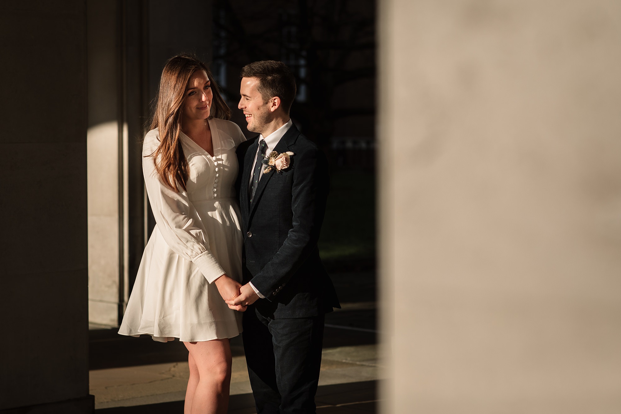 A couple stands holding hands, smiling at each other in front of the Hertford Register Office. The woman is in a white dress, and the man dons a dark suit with a boutonnière. They are bathed in warm sunlight, framed by the buildings stately stone columns.