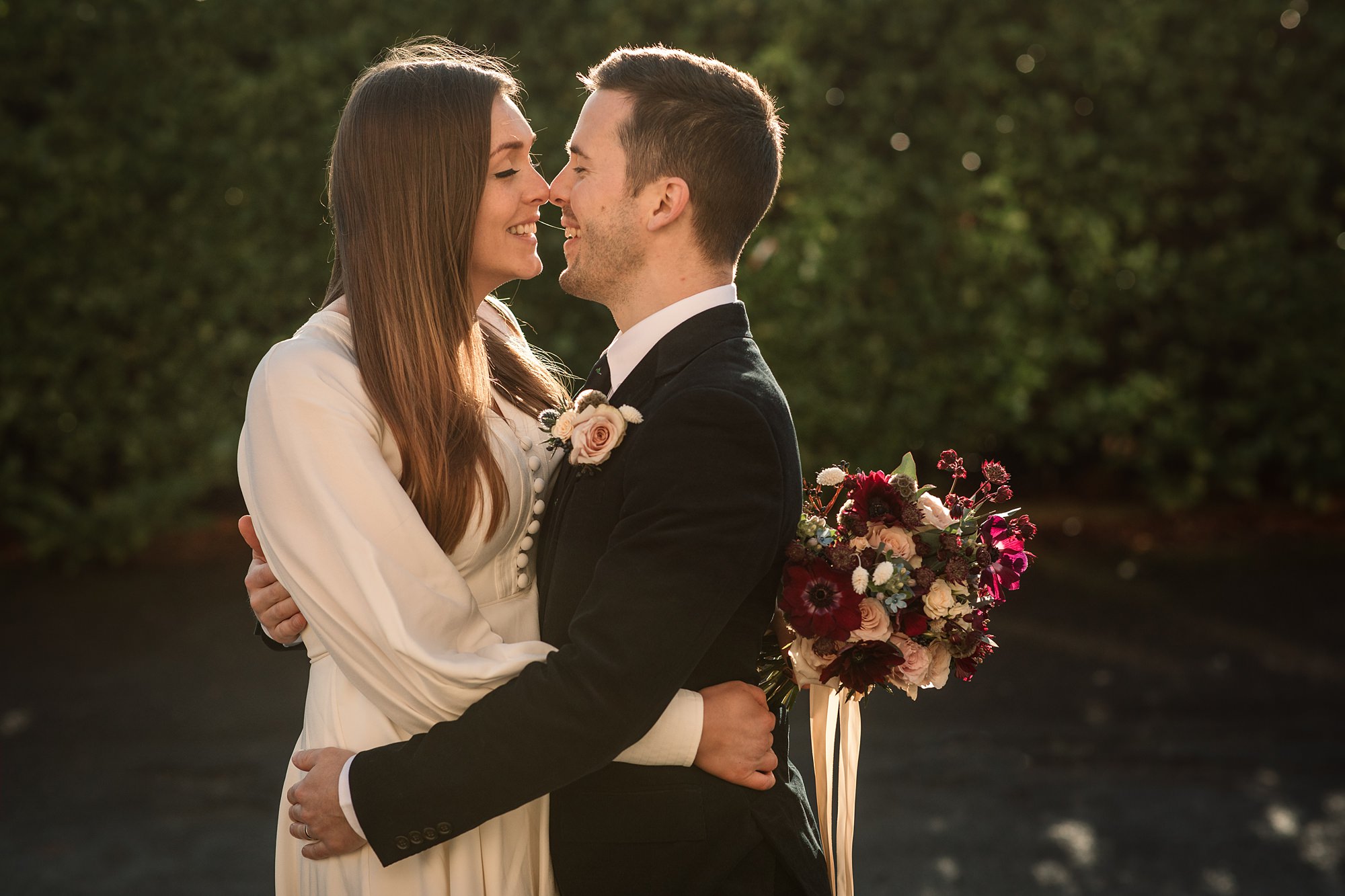 A couple embraces and smiles at each other outdoors, near the Hertford Register Office. The woman is in a white dress, the man in a suit with a boutonniere. She holds a bouquet of red and white flowers amidst lush greenery, all warmly lit by the setting sun.