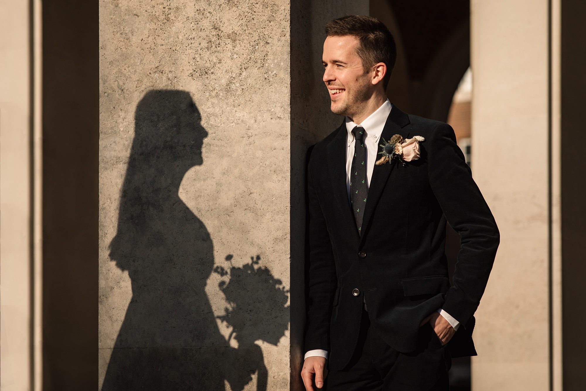A man in a suit smiles while standing next to a wall at Hertford Register Office. The shadow of a woman holding a bouquet is cast beside him, capturing a joyful moment from their wedding day.