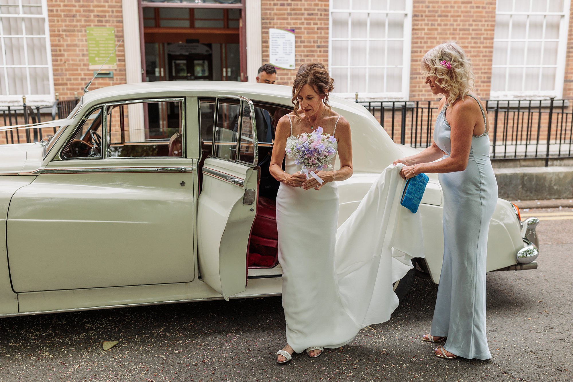 A bride in a white dress holding a bouquet steps out of a vintage white car at Hertford Register Office. Another woman in a blue dress holds the brides train, perfectly complementing the buildings brick walls and large windows.