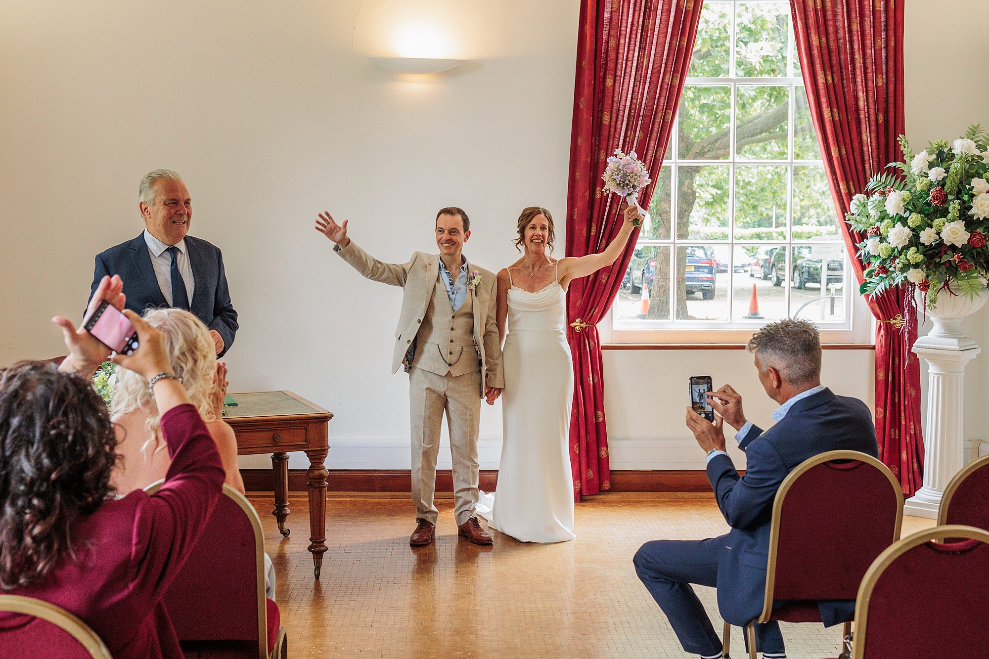 A newlywed couple celebrates joyfully at Hertford Register Office, waving to guests in a brightly lit room. The bride holds a bouquet, and the groom wears a beige suit. Guests sit in red chairs, applauding and taking photos by a large window with striking red curtains in the background.