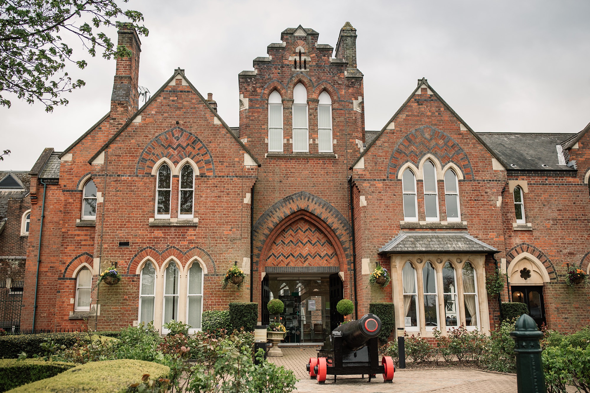 A Victorian-style red brick building, the St Albans Registry Office boasts tall arched windows and intricate brickwork at its entrance. In front, a black cannon on a red cart adds historic charm, set amidst lush greenery under a cloudy sky.