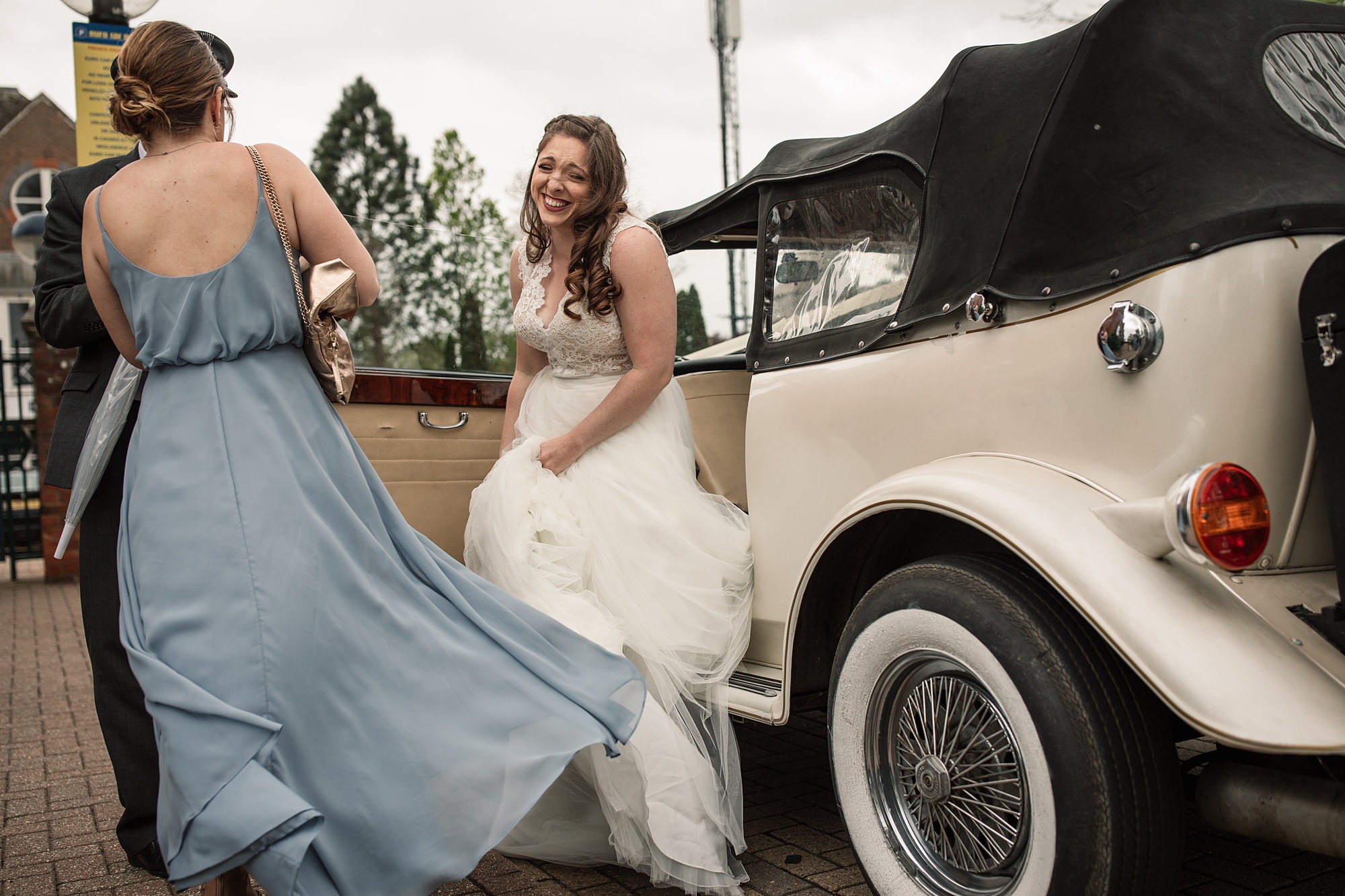A bride with long brown hair and a lace wedding dress steps out of a vintage car, smiling joyfully outside the St Albans Registry Office. An attendant in a blue dress holds the door open on the brick pavement. Trees and buildings grace the backdrop.