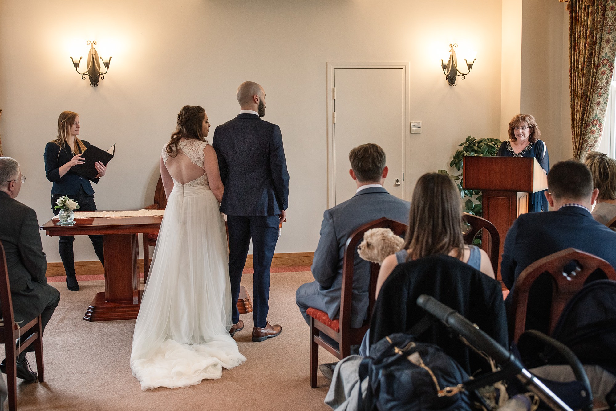 A bride and groom stand facing a woman officiating their wedding in the intimate St Albans Registry Office. Guests, some in formal attire, sit in rows. A flower arrangement graces a table beside the couple. Sconces and curtains adorn the walls while a small dog sits among the guests.
