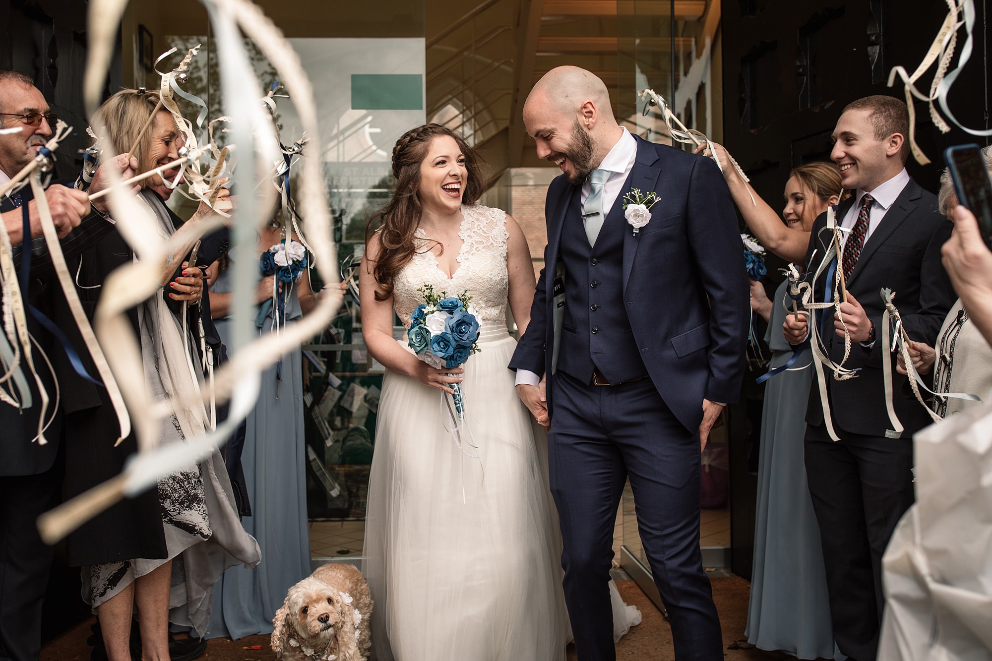 A bride and groom, elegantly dressed, smile as they exit the St Albans Registry Office, surrounded by joyful guests waving streamers. The bride holds a bouquet of blue flowers, with a small dog playfully trotting at their feet.