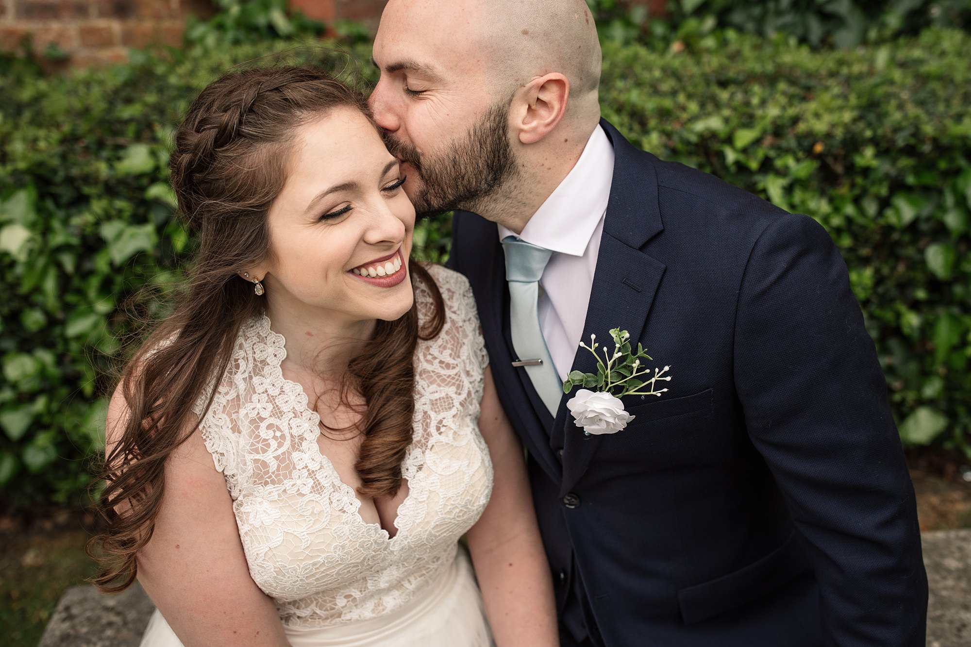 A bride in a lace dress smiles as the groom, in a navy suit with a white boutonniere, kisses her forehead. Theyre outdoors amid the lush greenery near St Albans Registry Office, capturing a moment of pure love and joy.