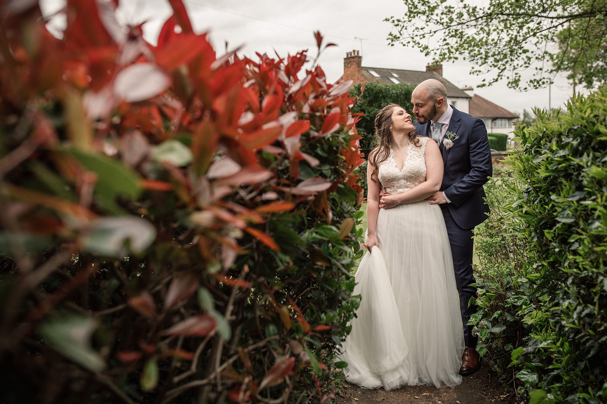 A bride in a white gown stands with a groom in a navy suit, surrounded by lush greenery and vibrant red leaves at the St Albans Registry office. The groom gently kisses the brides forehead, with a charming house visible in the background.