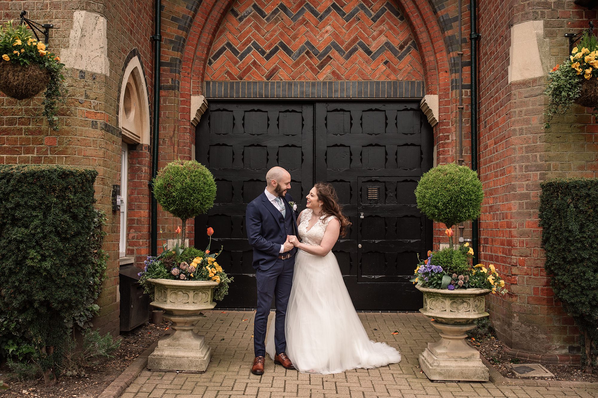 A bride and groom stand beaming in front of the large black door at St Albans Registry office, set in a brick wall with zig-zag patterns. They are surrounded by potted plants and greenery, including topiary trees and colorful flowers.
