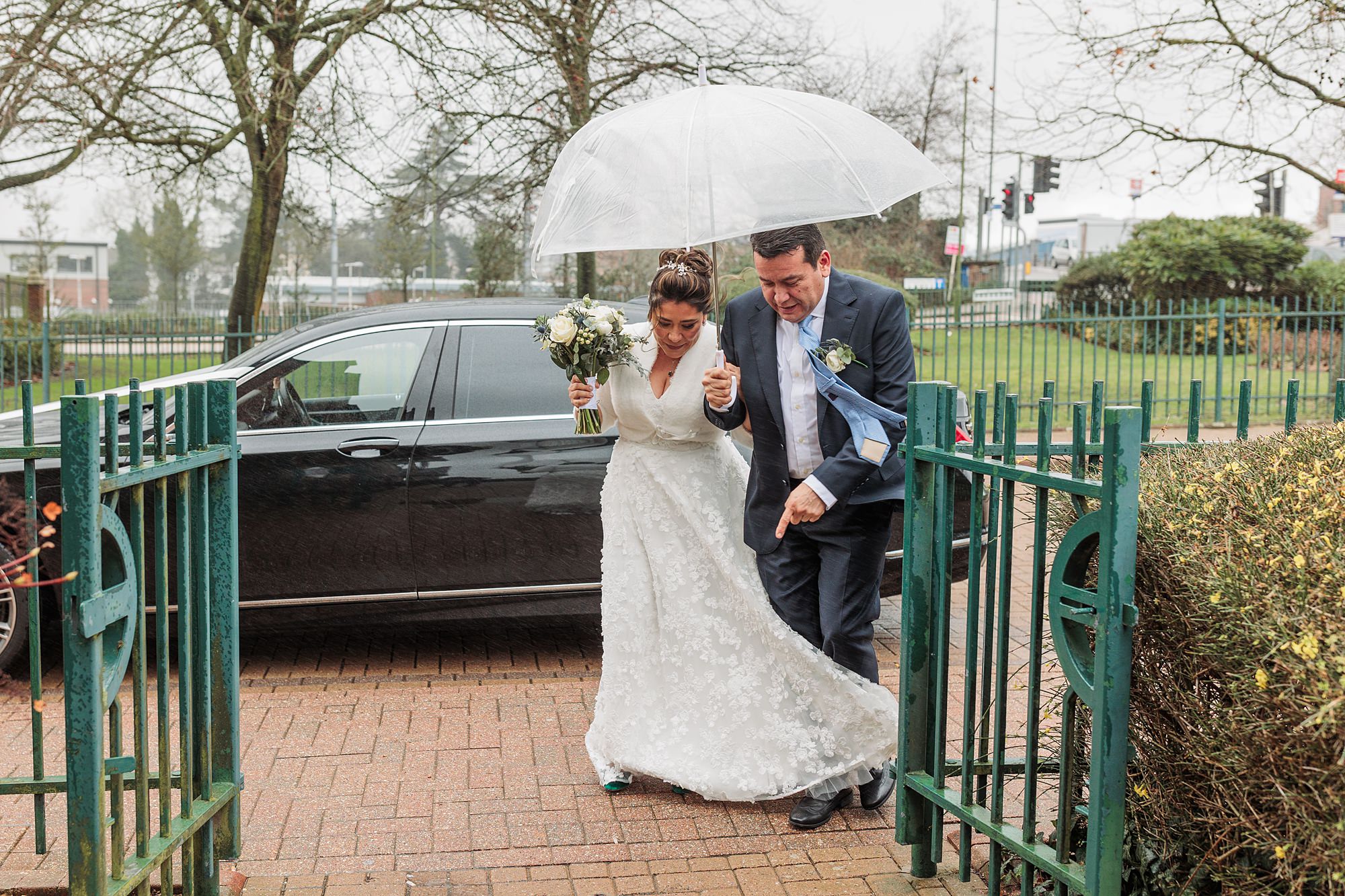 A bride in a white wedding dress, holding a bouquet, walks under a clear umbrella with a man in a suit. They enter the green gate of St Albans Registry Office on a rainy day, with trees and a parked car in the background.