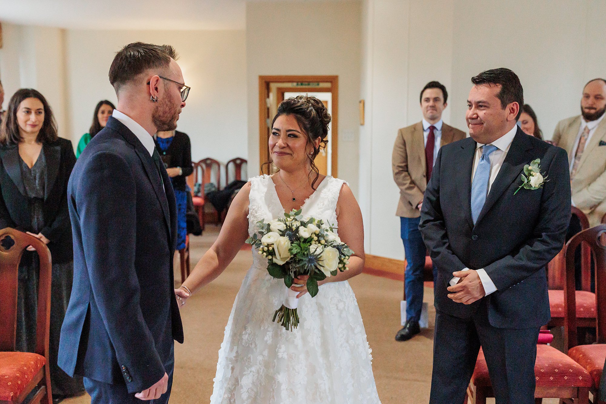 A bride in a white dress, holding a bouquet, stands smiling at the groom in the elegant St Albans Registry Office. The groom is beside a man in a suit with a flower boutonniere. They are surrounded by chairs and well-wishers in the background.
