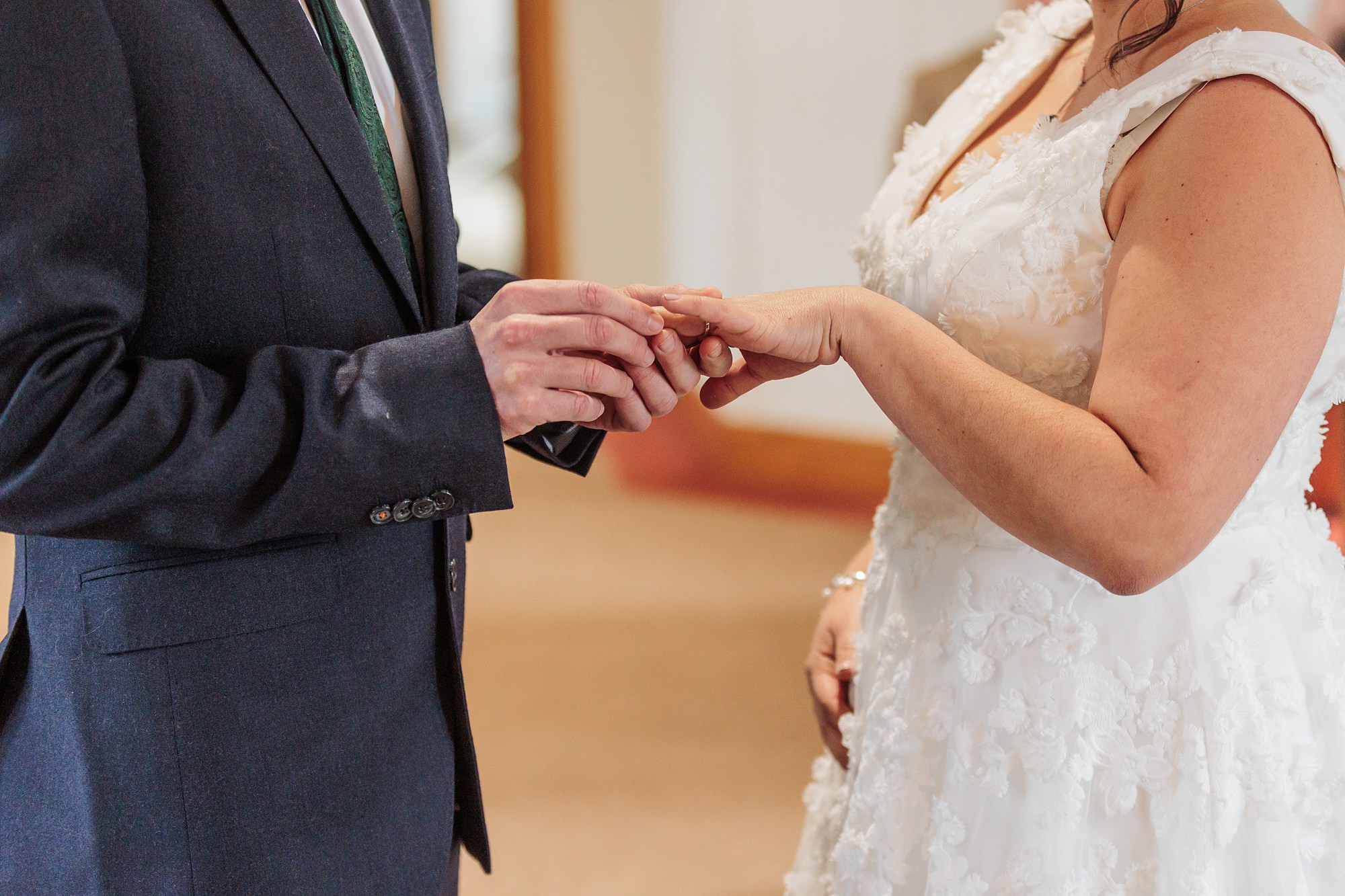 At the St Albans Registry Office, a bride and groom exchange rings during their wedding ceremony. The groom, dressed in a dark suit, gently places a ring on the brides finger as she stands elegantly in her white lace gown. The focus is on this heartwarming moment of union.