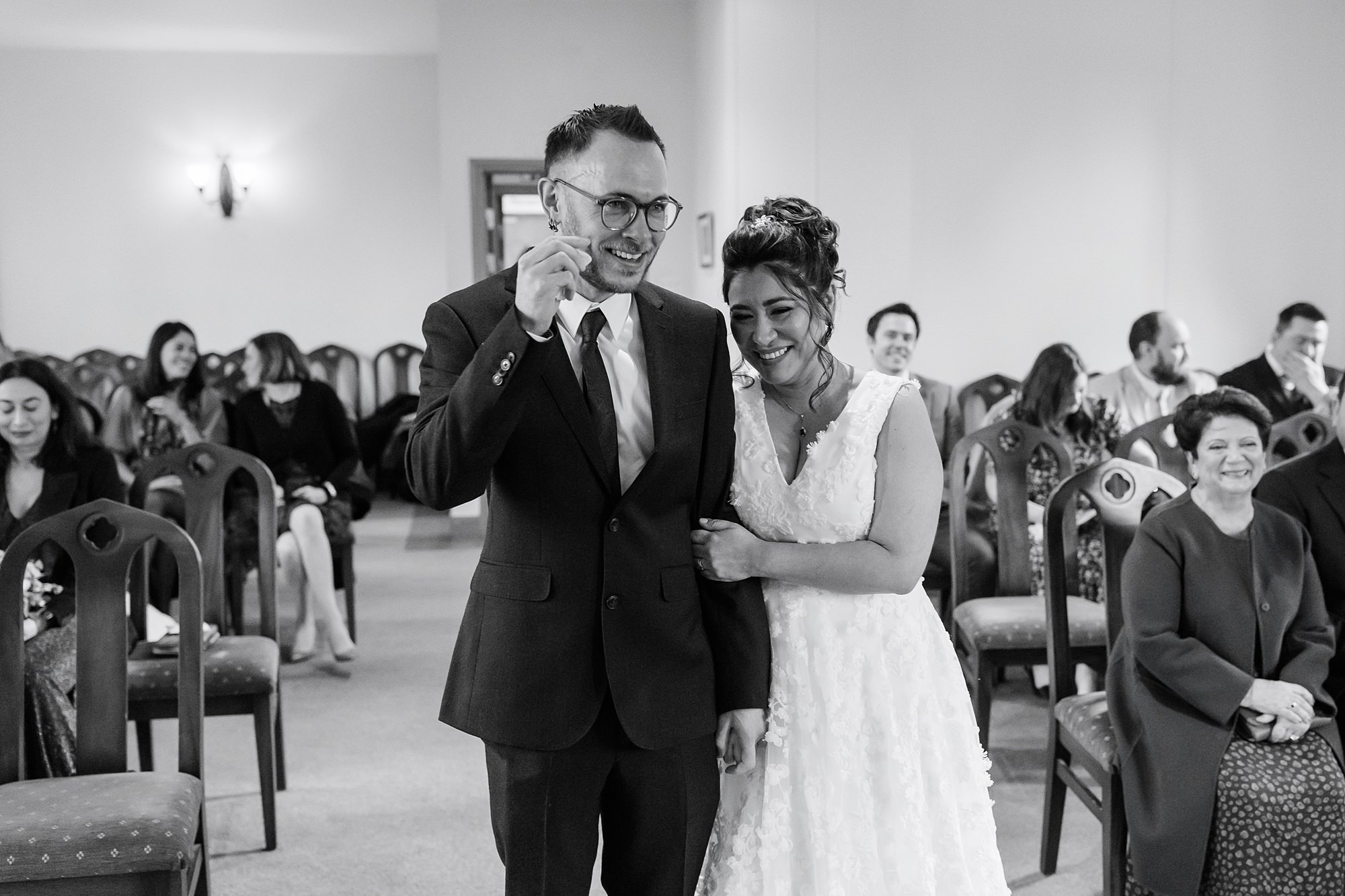 A bride and groom, both smiling and teary-eyed, stand arm in arm at their St Albans Registry office ceremony. Guests seated in the background watch the couple with warm smiles. The scene is intimate and joyful.