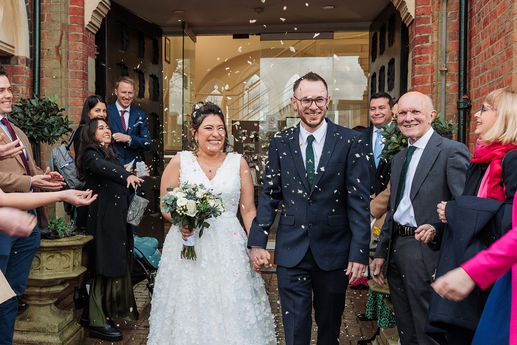A bride in a floral white dress and a groom in a navy suit with a green tie walk hand in hand under a shower of confetti. They are surrounded by smiling guests outside a brick building. The bride holds a bouquet and both are beaming.