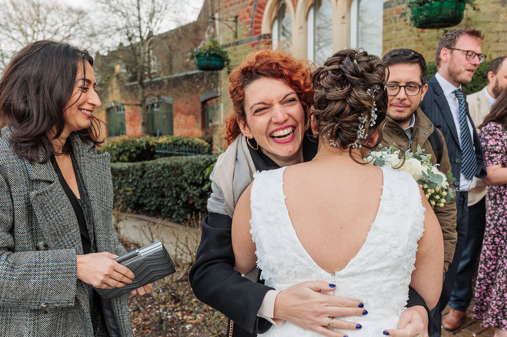 Outside the St Albans Registry Office, a bride in a white dress with a floral pattern embraces a laughing woman with curly red hair. Surrounded by several people near the charming brick building and lush greenery, the atmosphere is joyous and celebratory.