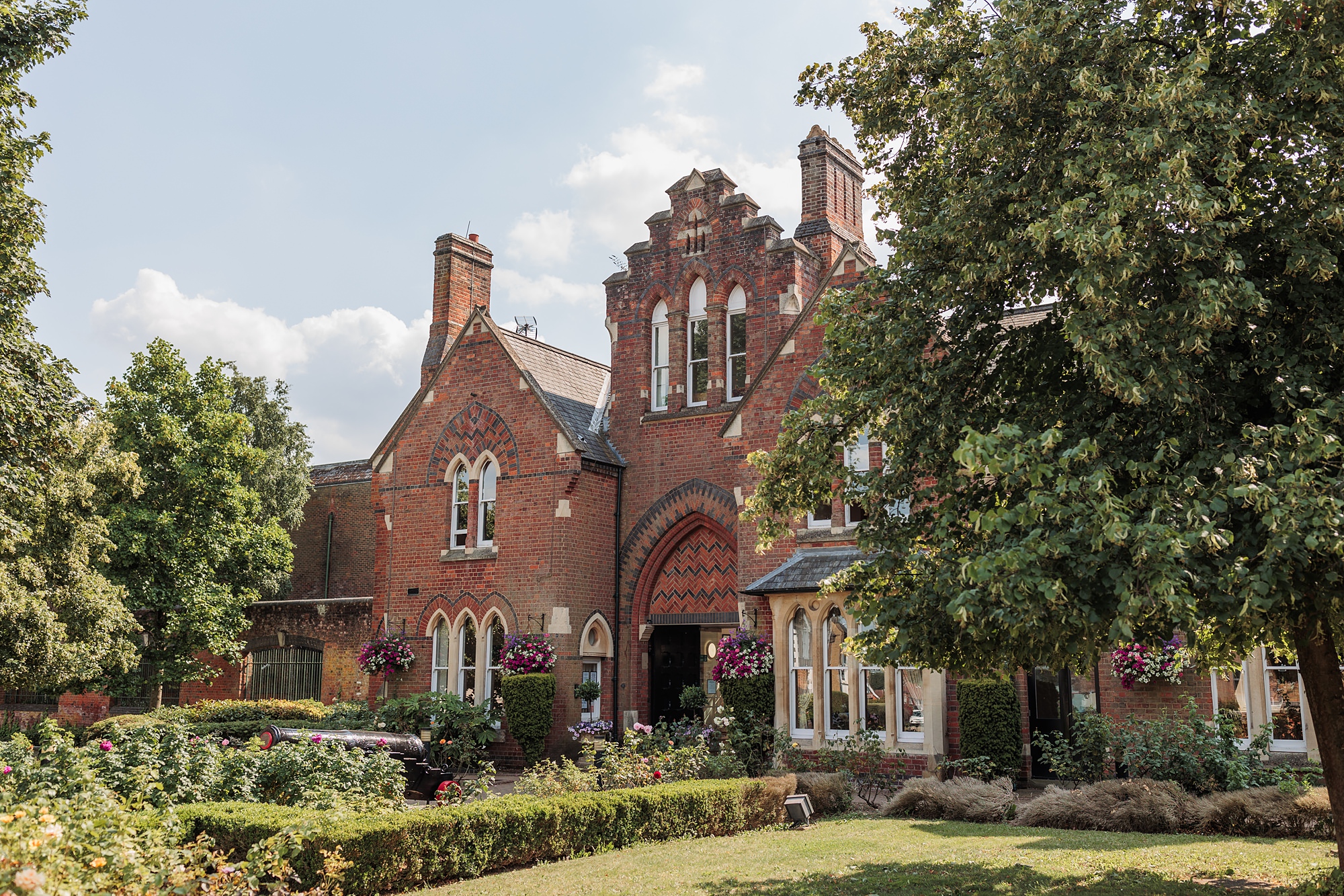 A charming red-brick Victorian building with intricate architectural details and arched windows, surrounded by lush greenery and flowering plants, serves as the St Albans Registry Office. A large tree partially shades the front of this picturesque setting.
