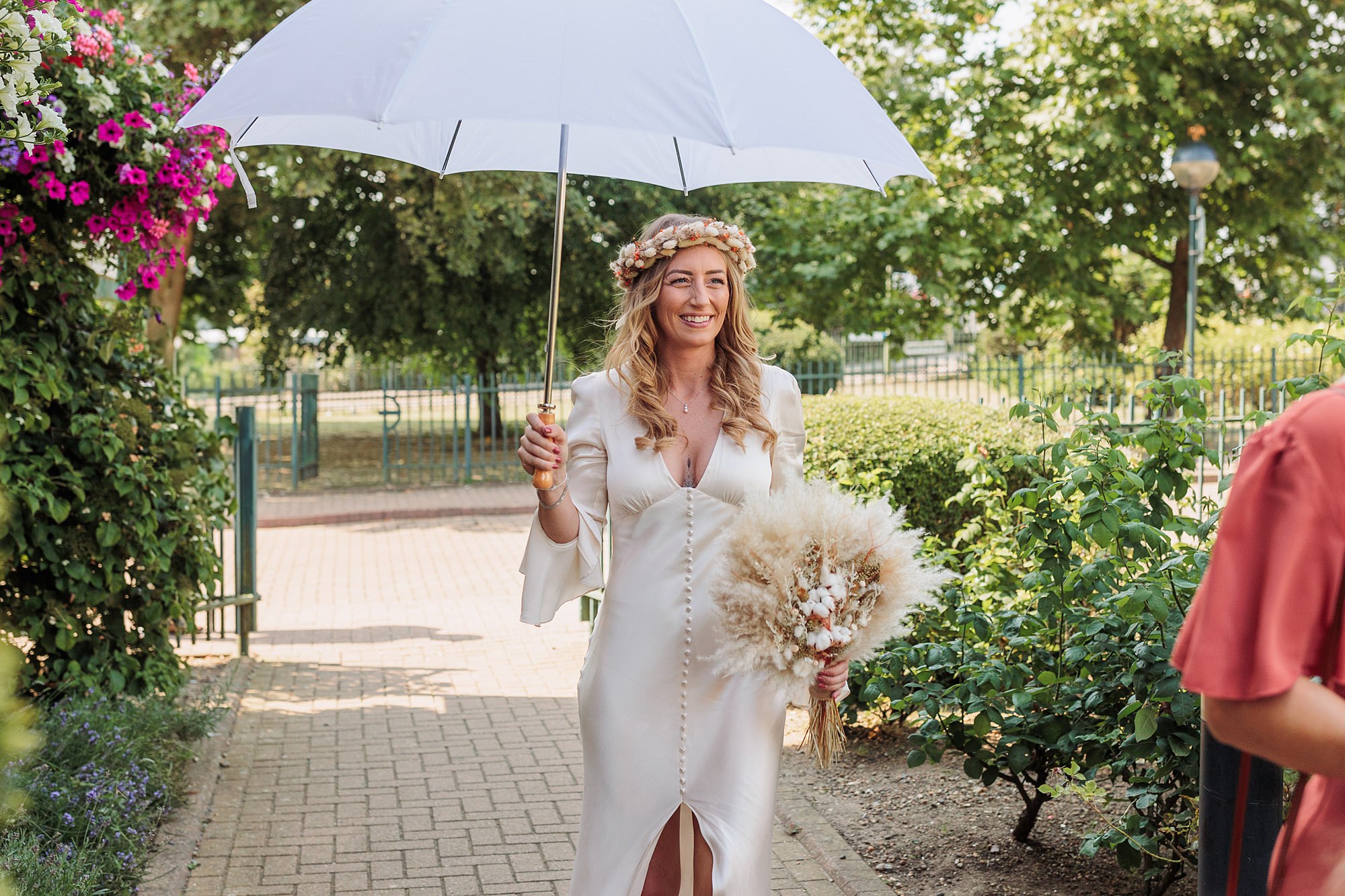 A smiling bride in a white dress holds a large, white umbrella and a bouquet of dried flowers as she walks near St Albans Registry Office. Adorned with a floral crown, she strolls along a sunlit path surrounded by greenery.