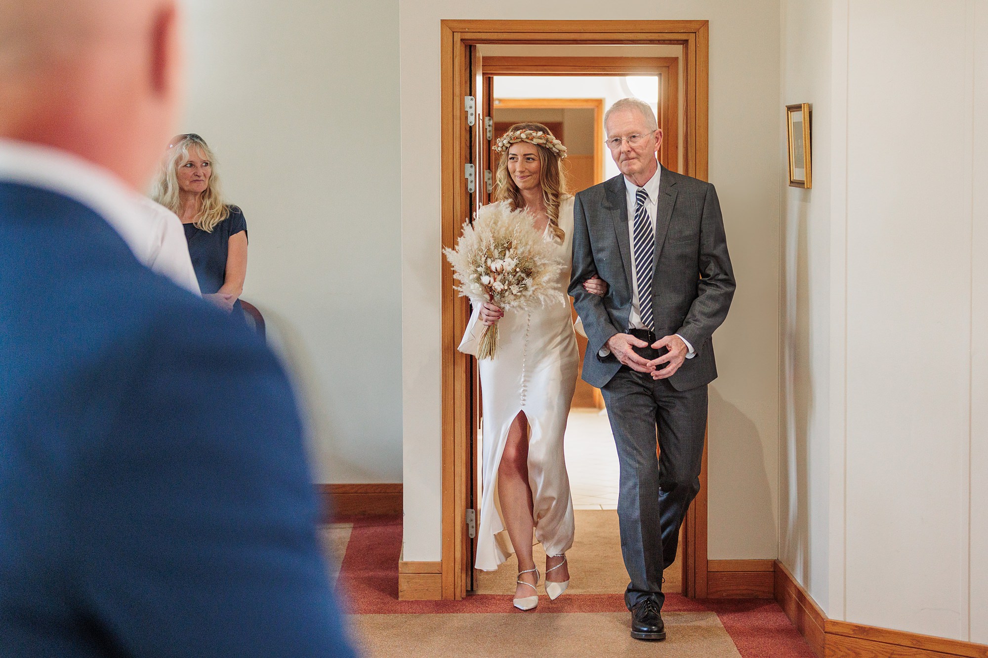 A bride in a white dress and floral crown walks down the aisle of St Albans Registry Office with an older man in a suit, holding a bouquet of dried flowers. They enter through a wooden doorway as a man in a blue suit looks on.