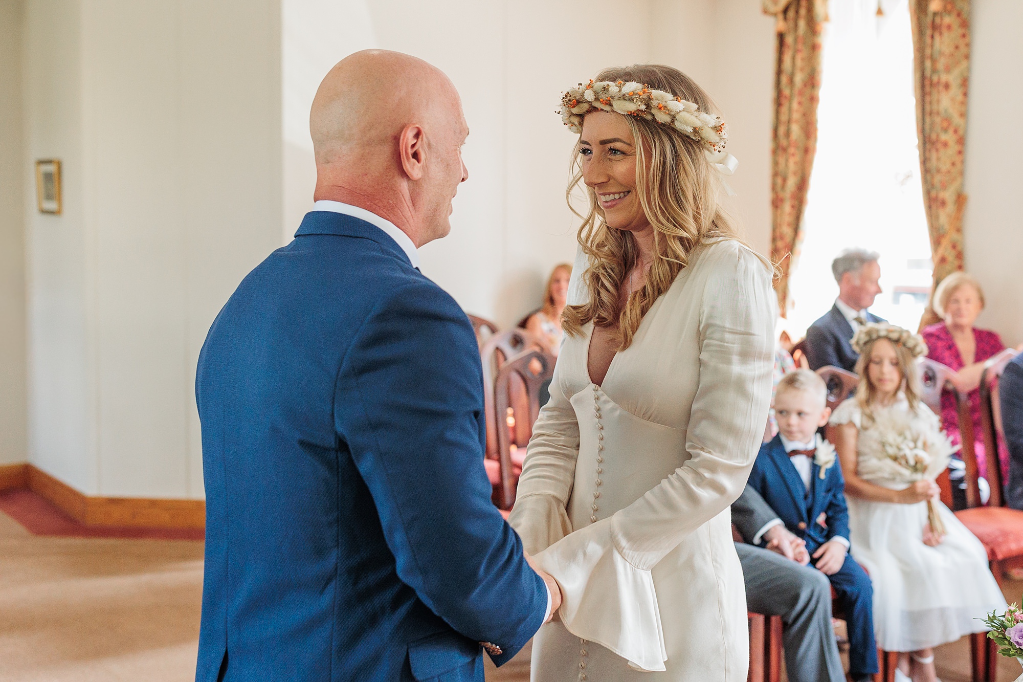 A couple stands facing each other, holding hands at St Albans Registry Office during their wedding ceremony. The woman wears a white dress and a floral crown, while the man is in a blue suit. Guests are seated in the background, witnessing their vows.
