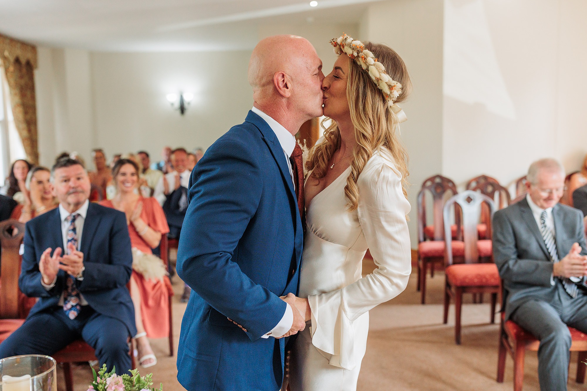 At the St Albans Registry office, a bride and groom share a tender kiss during their wedding ceremony. The groom looks dapper in a blue suit, while the bride dazzles in her white dress and floral headband. Guests are seated in the background, clapping and smiling warmly.