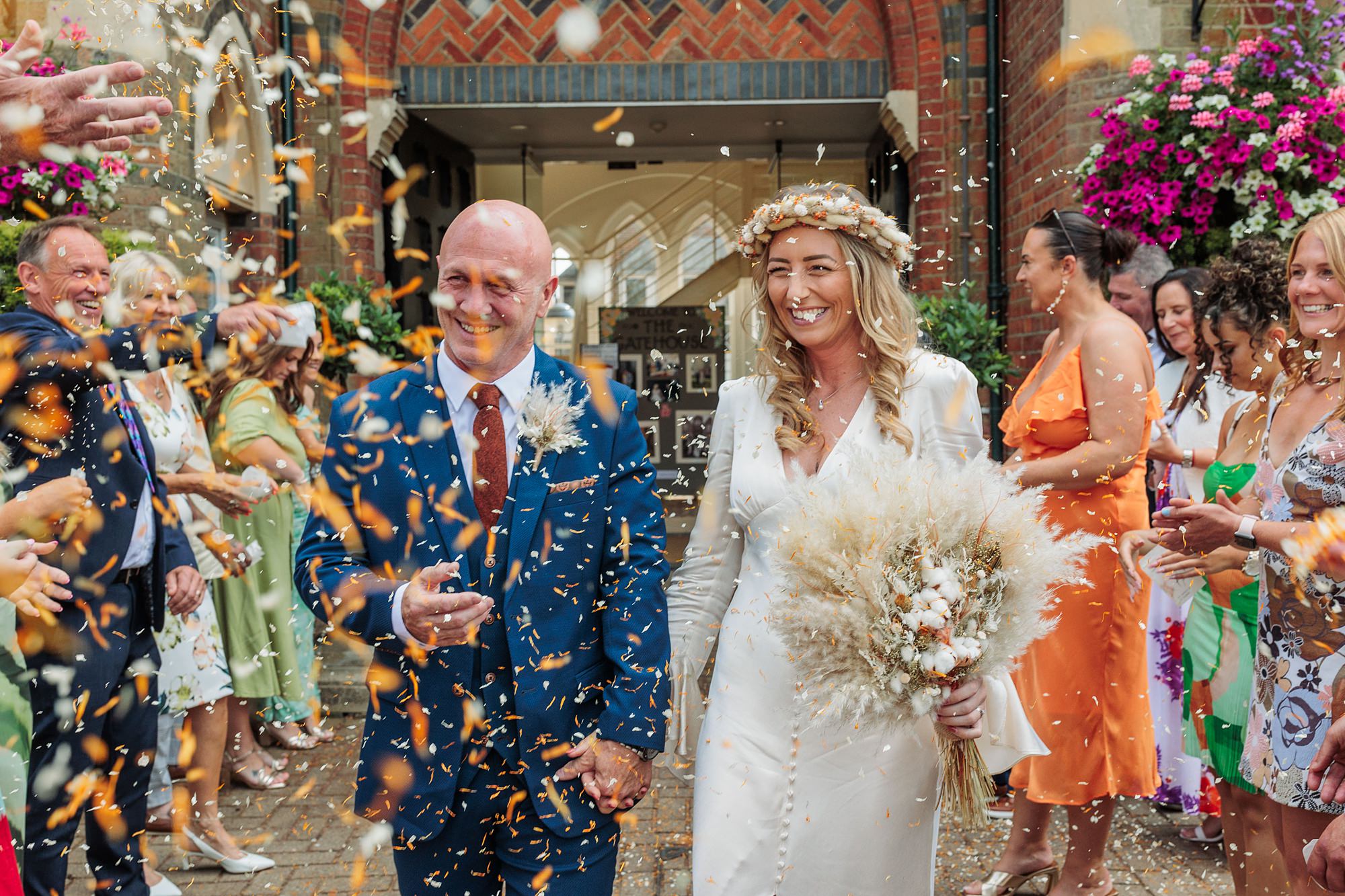 The bride and groom beam as they walk down the aisle at St Albans Registry office, guests showering them with colorful confetti. She carries a bouquet of dried flowers, her floral crown perfectly complementing his blue suit. Brick architecture and hanging flower baskets add to the charm.