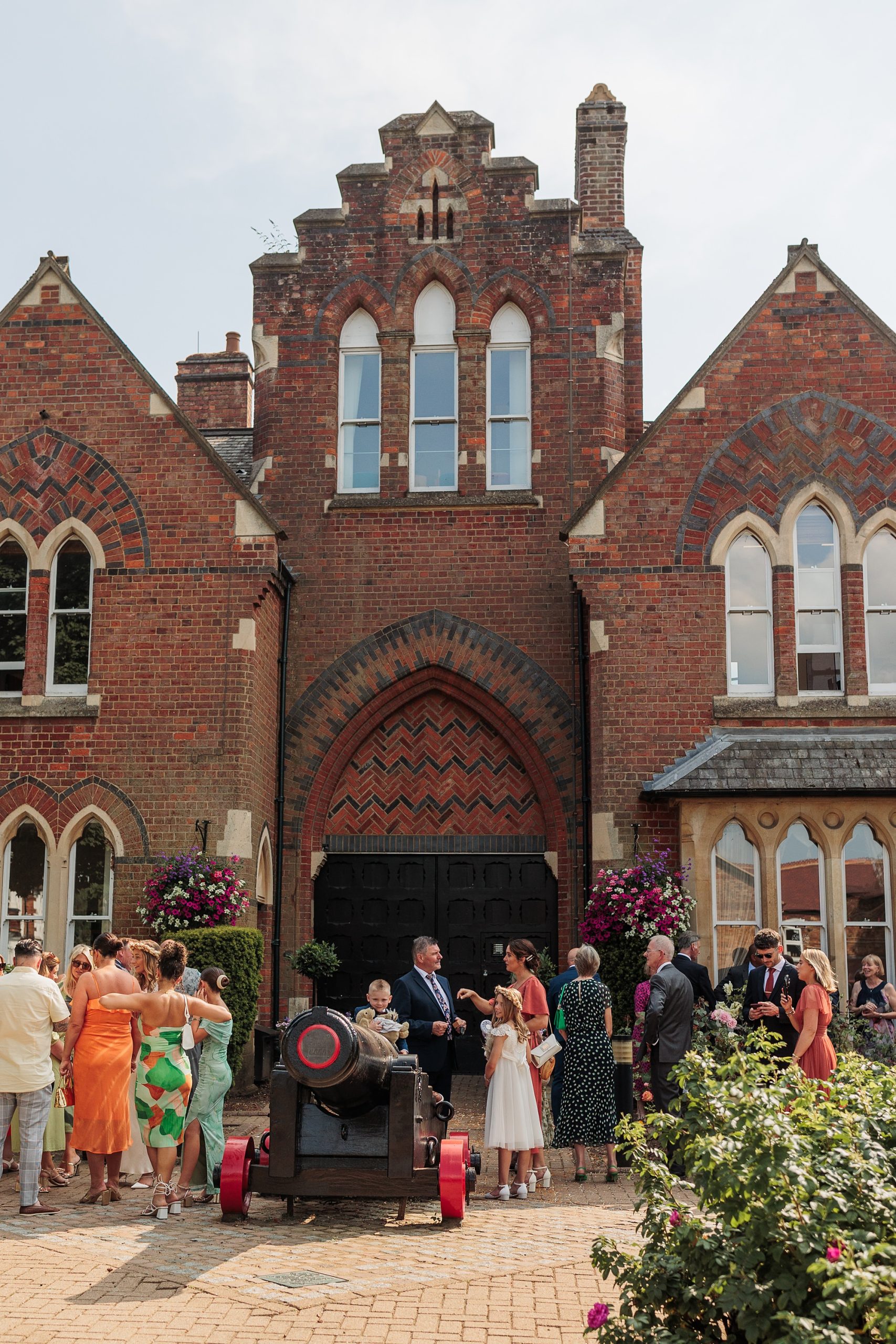 A lively group of people gather in front of the historic St Albans Registry Office, a beautiful brick building with gothic-style architecture. Some are dressed in formal attire, chatting and smiling beside a decorative cannon and vibrant flower arrangements.